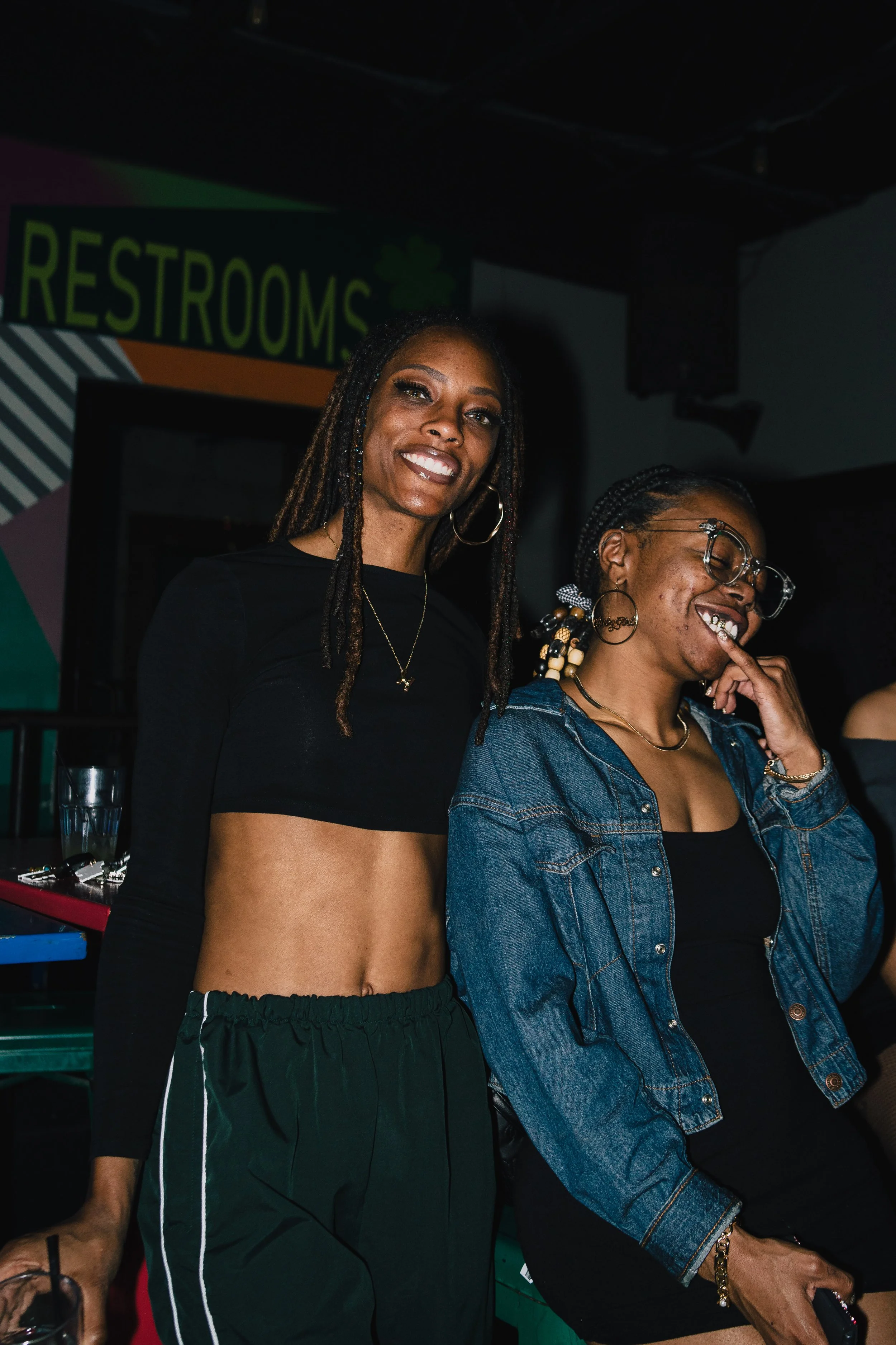 Two women smiling and laughing in a dimly lit room, with a sign that says 'Restrooms' in the background.