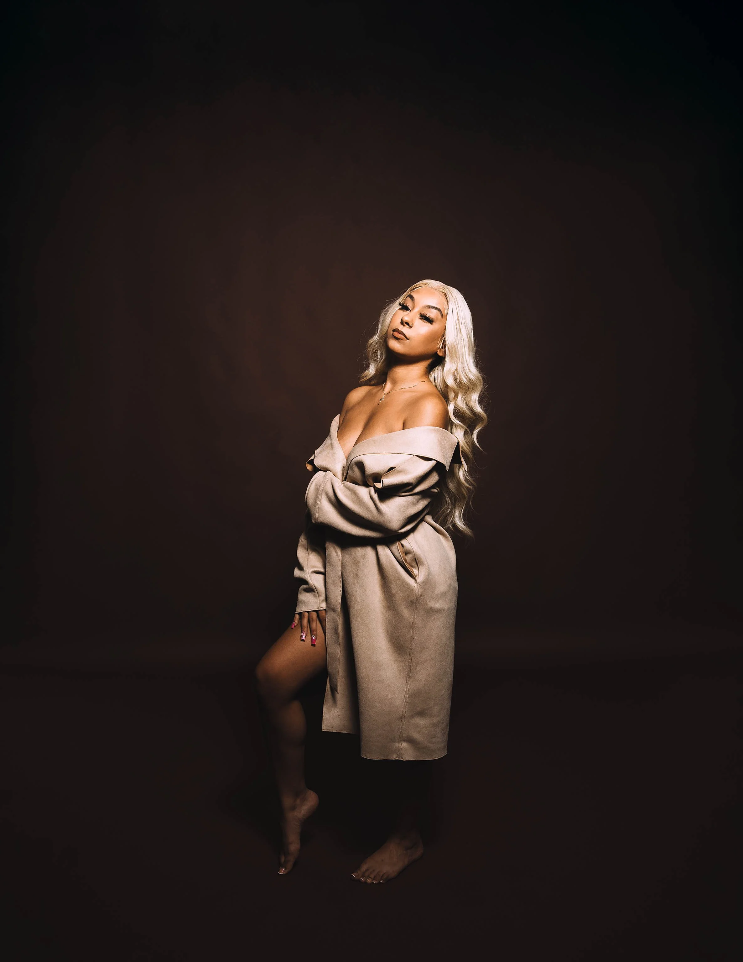 A woman with long, blonde, wavy hair posing in a beige trench coat against a dark background, standing on one foot with her arms crossed.