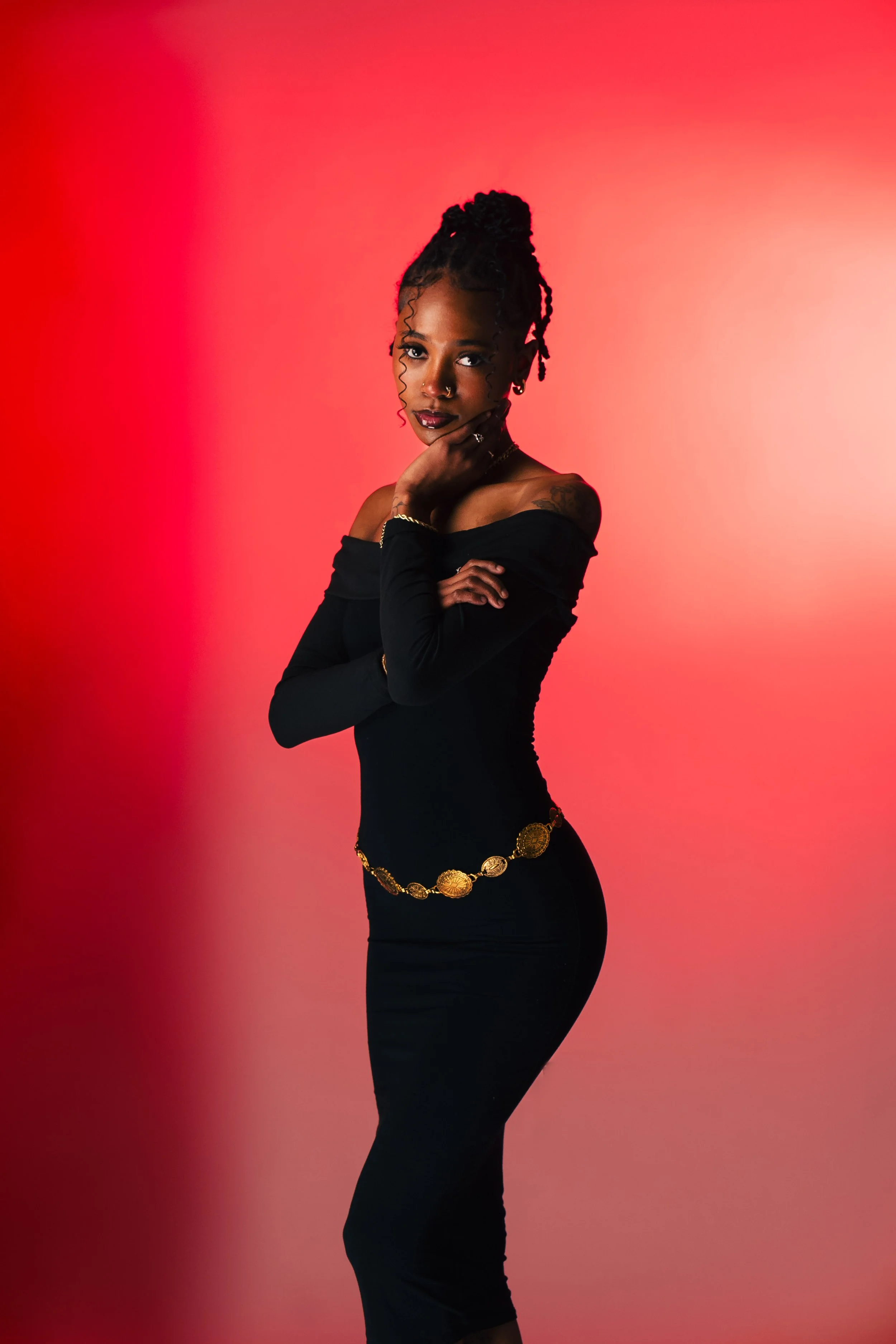 A woman with dark skin and styled bantu knots hair poses against a gradient red background, wearing a black off-shoulder dress with a gold belt, looking confidently into the camera with one hand near her face.