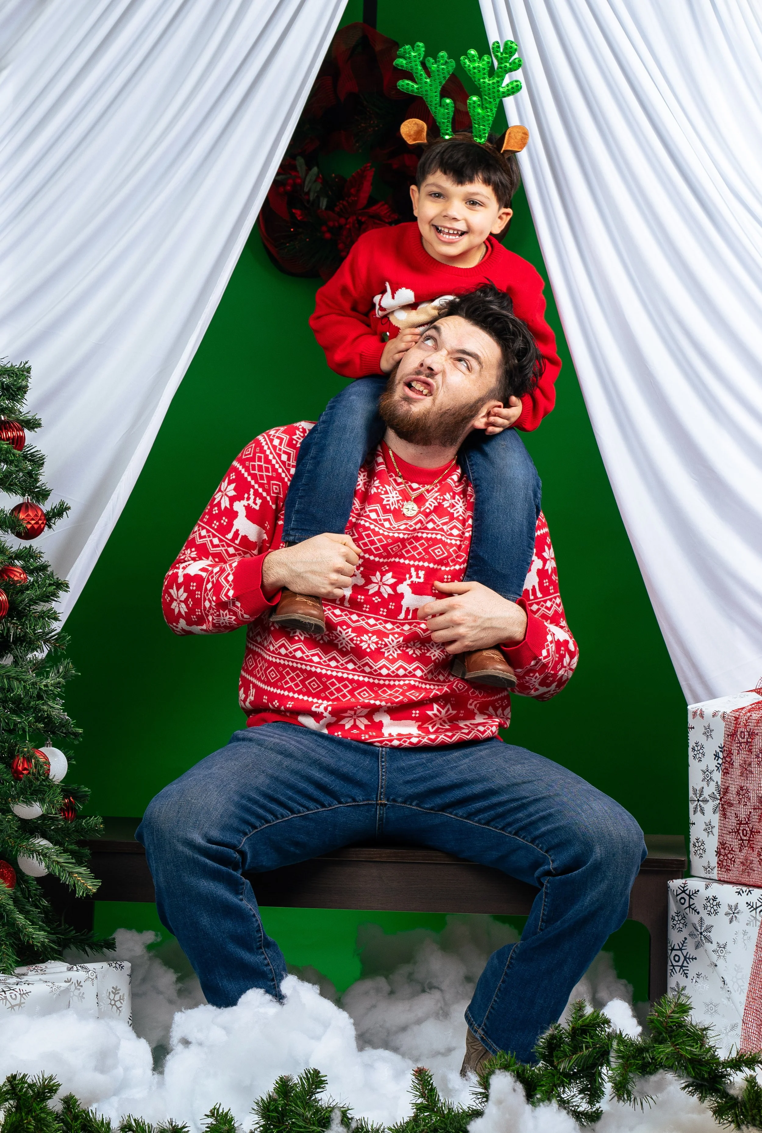 A man and a boy posing with Christmas decorations, the boy sitting on the man's shoulders, both wearing holiday sweaters. The boy has reindeer antlers on his head, and the man has facial expressions as if saying a funny line. Christmas tree, presents