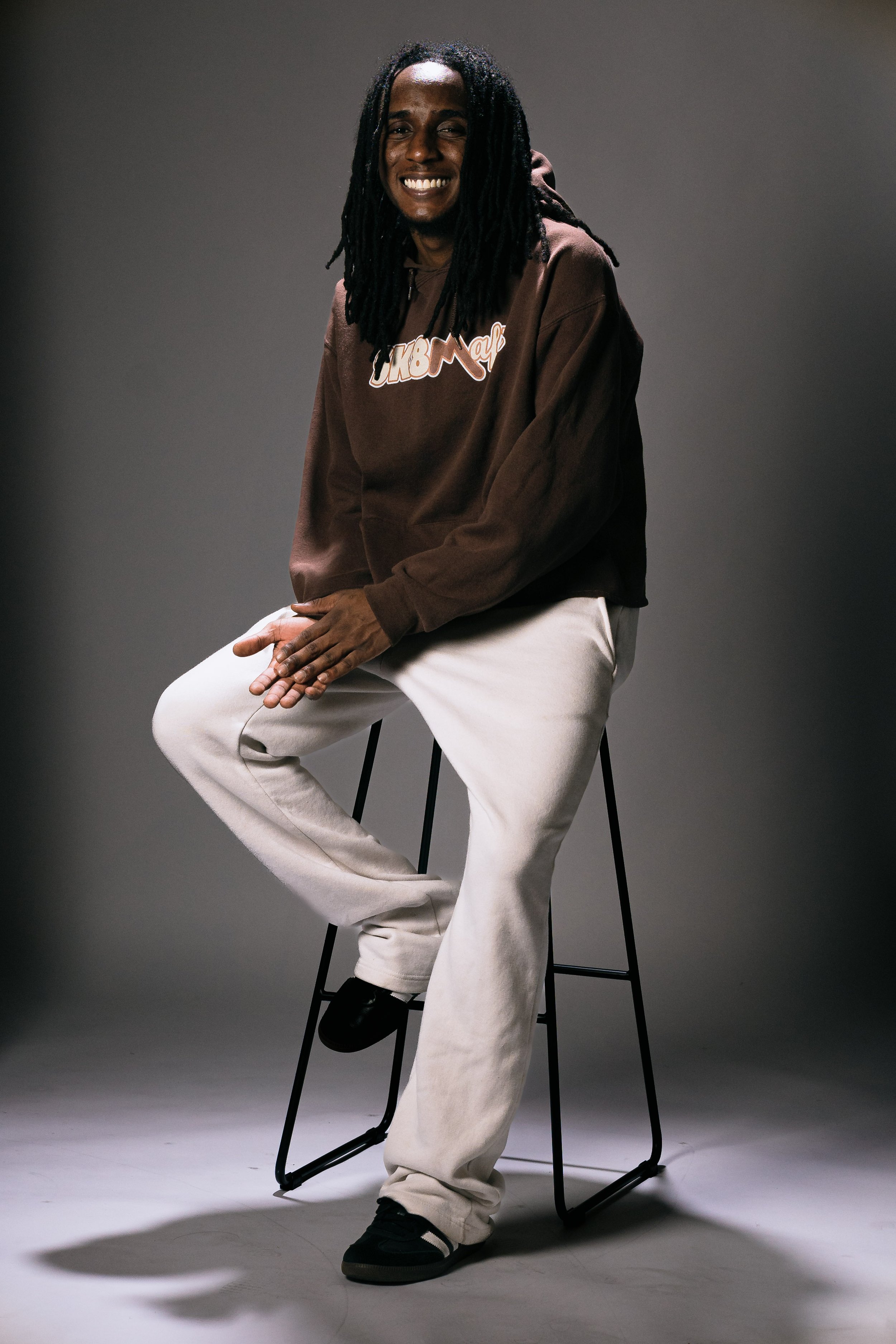 A man with long dreadlocks sitting on a black stool, smiling, wearing a brown hoodie and light-colored pants, against a plain dark background.