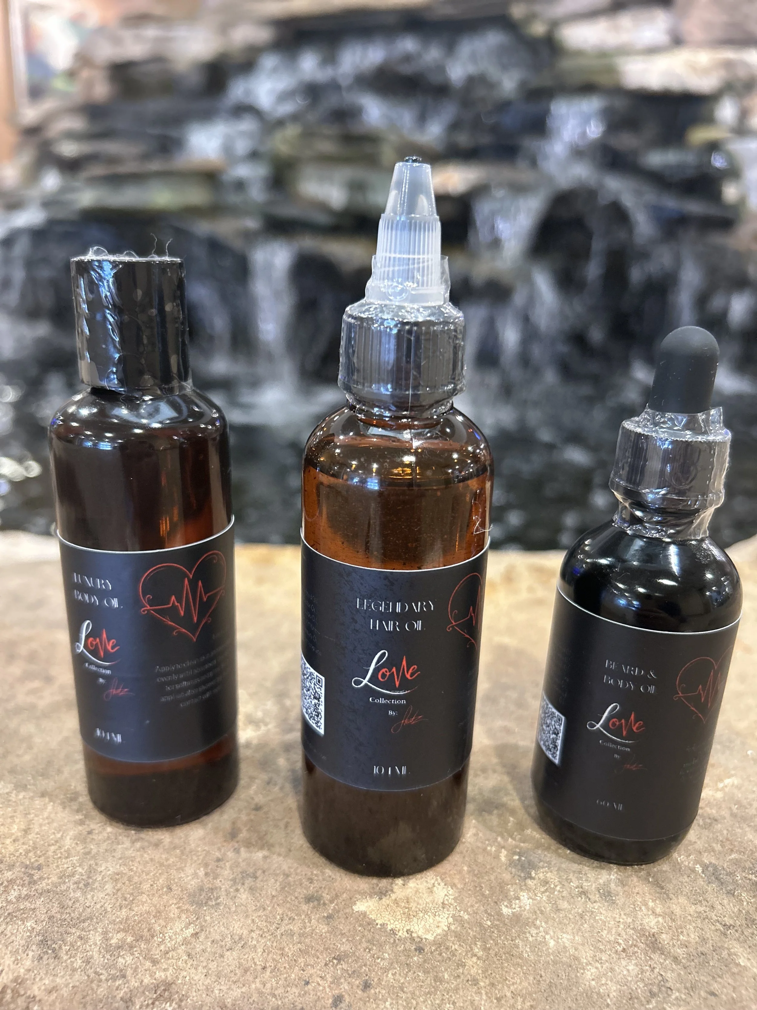 Three small bottles of body oil with black labels featuring red and white text, placed on a textured surface in front of a waterfall background.