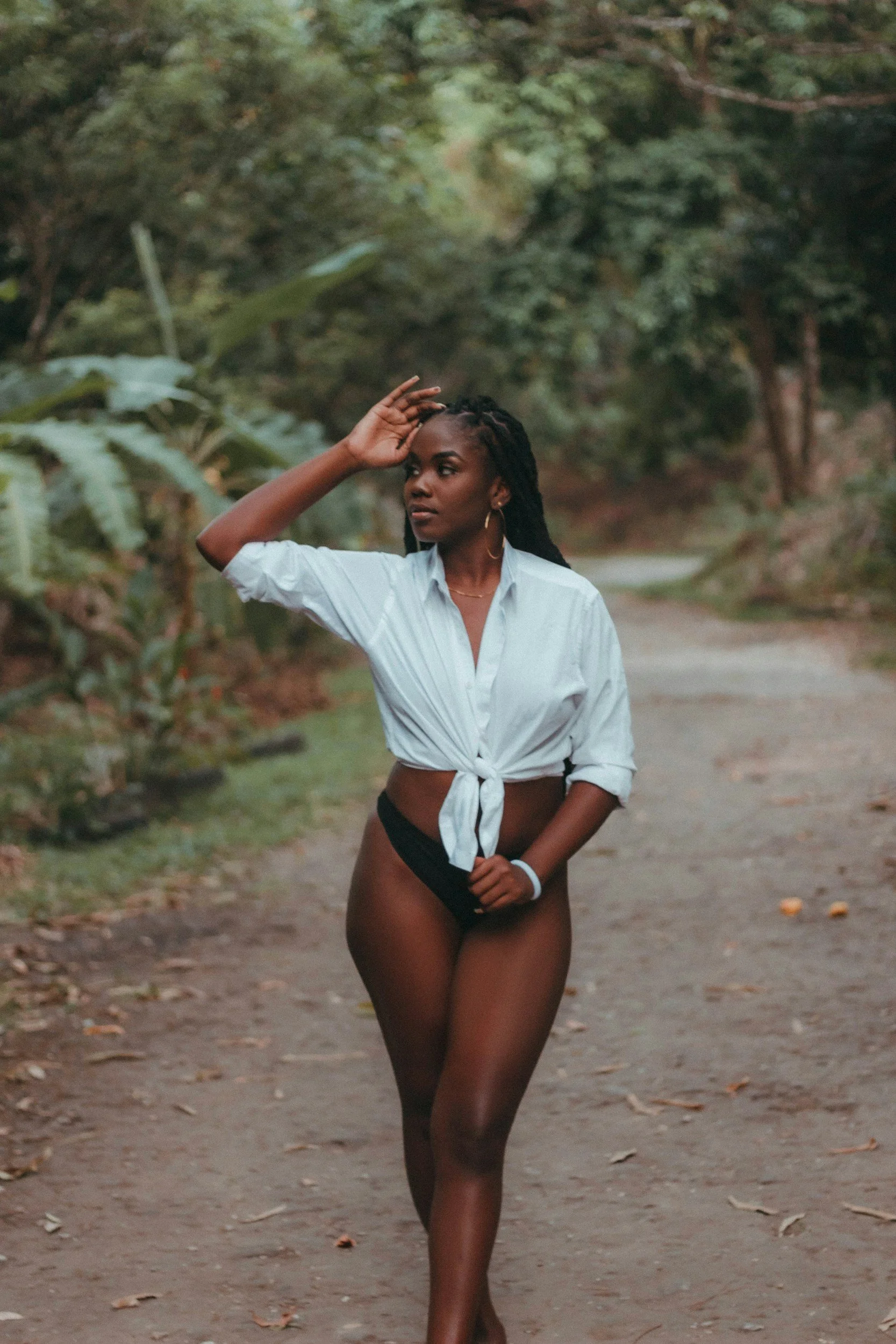 A woman with dark skin and long braids walks on a wooded trail, wearing a tied-up white shirt and black underwear, with her left hand near her forehead and her right hand holding her shirt.