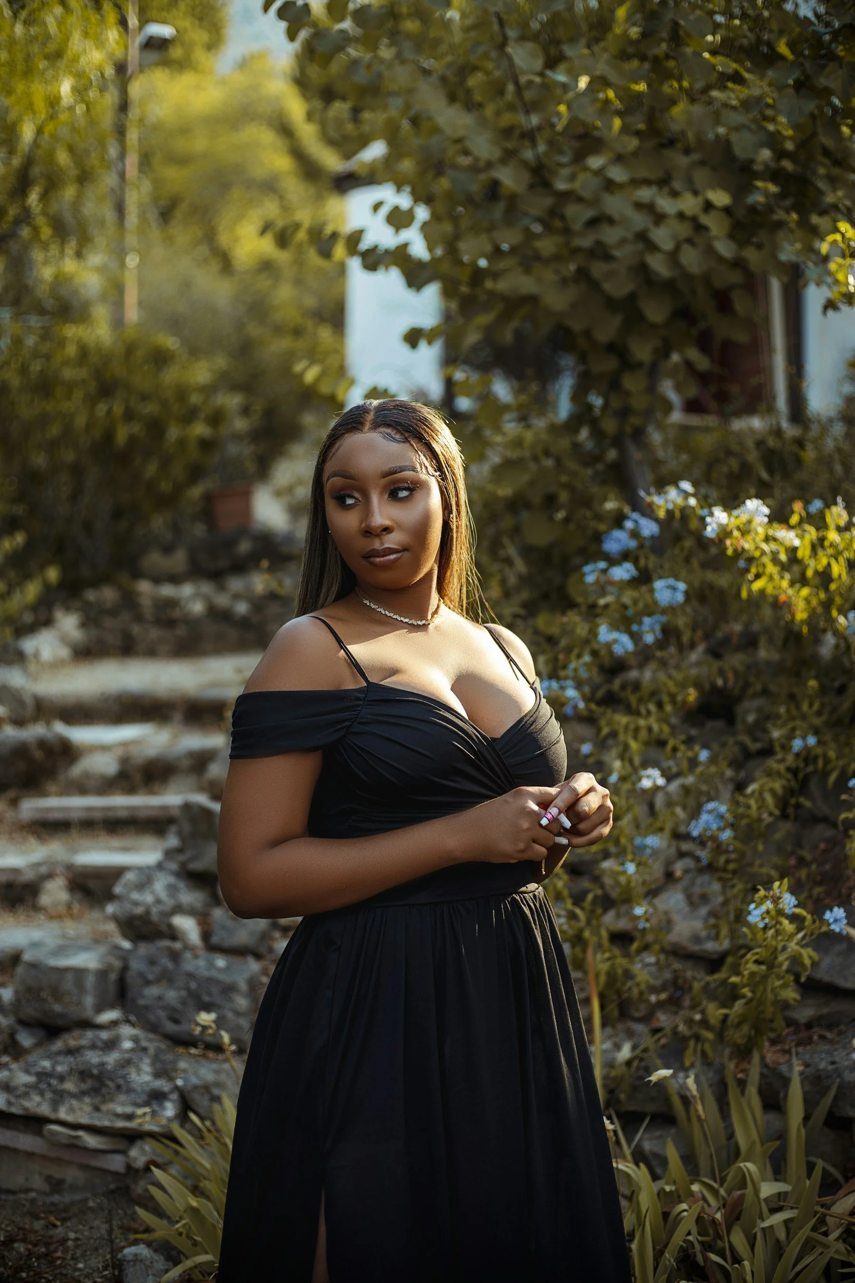 A young woman in a black off-shoulder dress standing outdoors amid green foliage and flowering plants, with sunlight illuminating her face and surroundings.