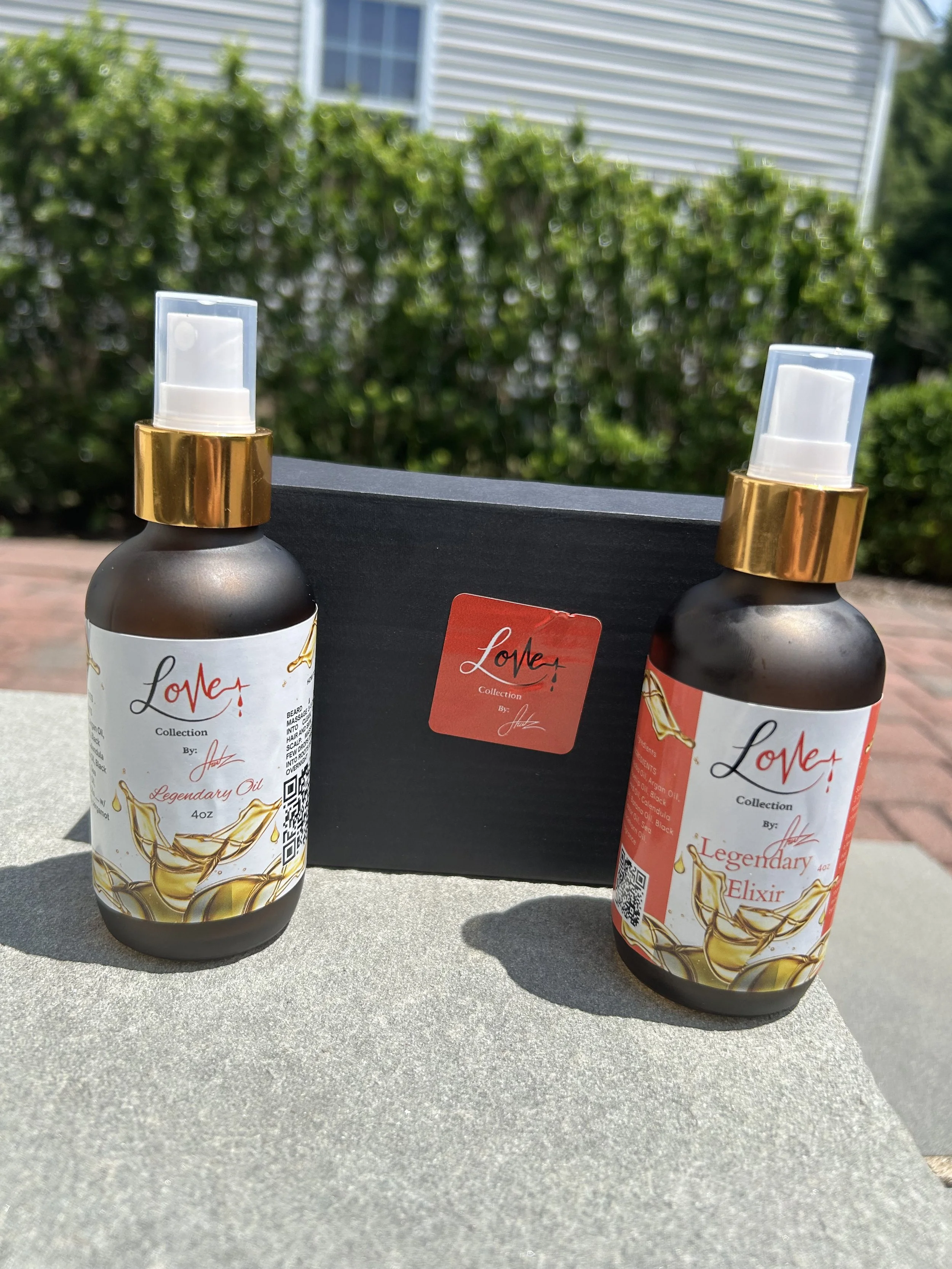 Two bottles of hair oil with a black box in the background, each with a gold cap and white pump, outdoor setting with greenery and a house in the background.