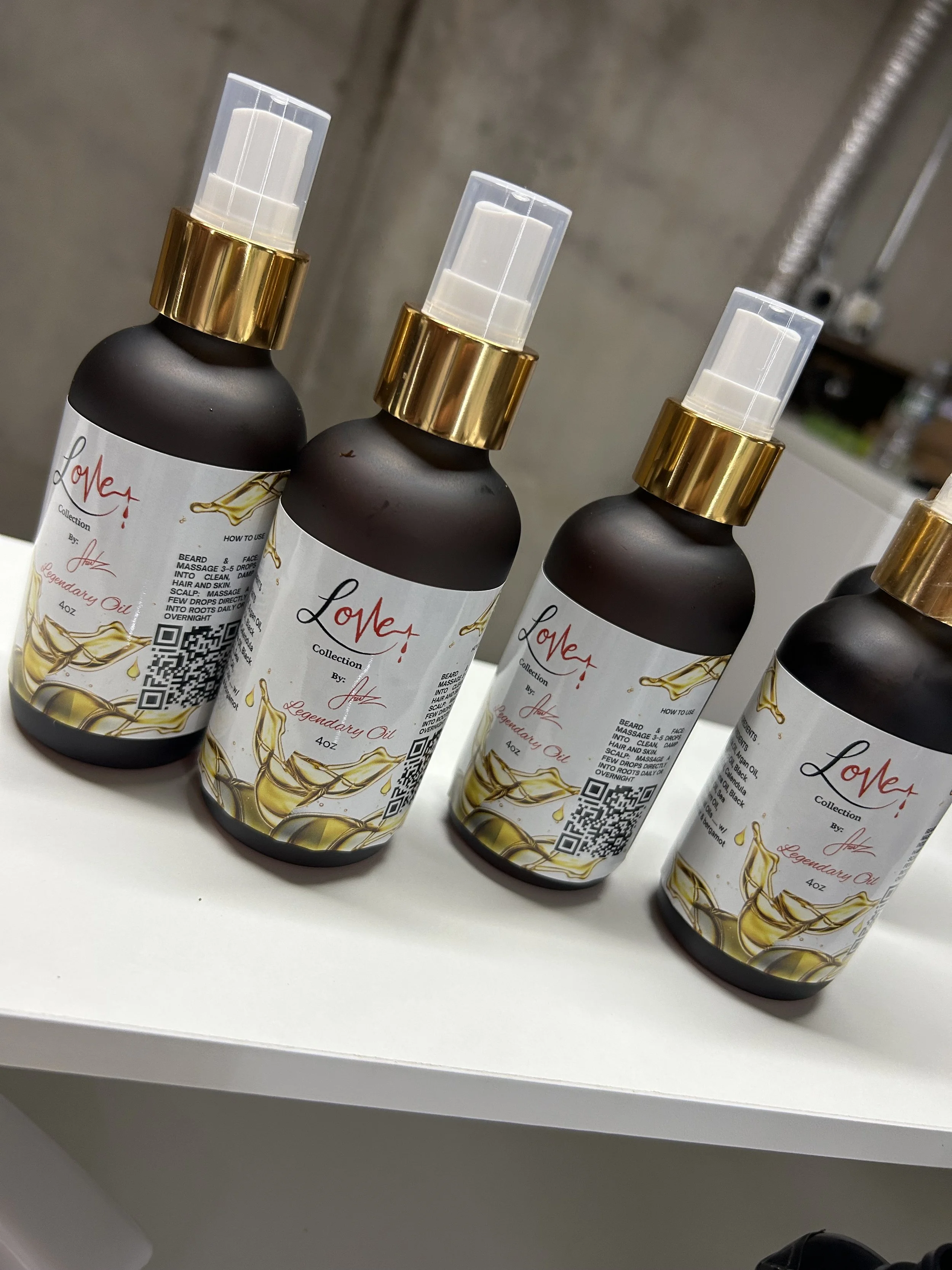 Three bottles of Love Legendary Oil spray with black matte finish, gold pump tops, and white labels with gold accents, arranged on a white surface in front of a concrete wall with visible pipe.
