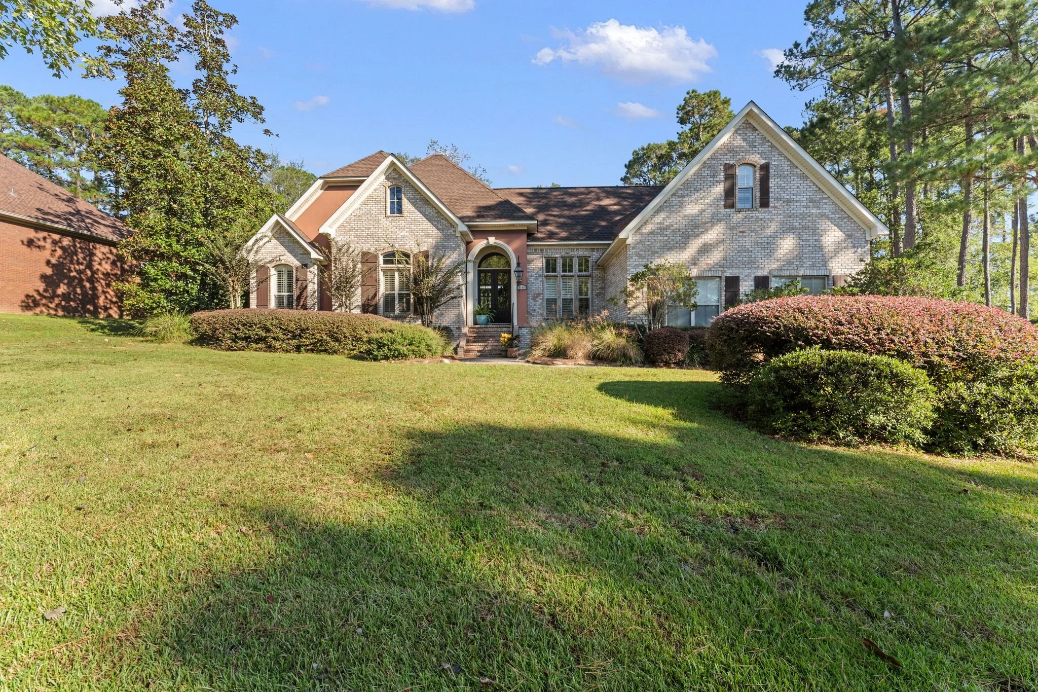 Front view of a large suburban house with a well-maintained lawn and shrubbery, trees in the background, and a blue sky.