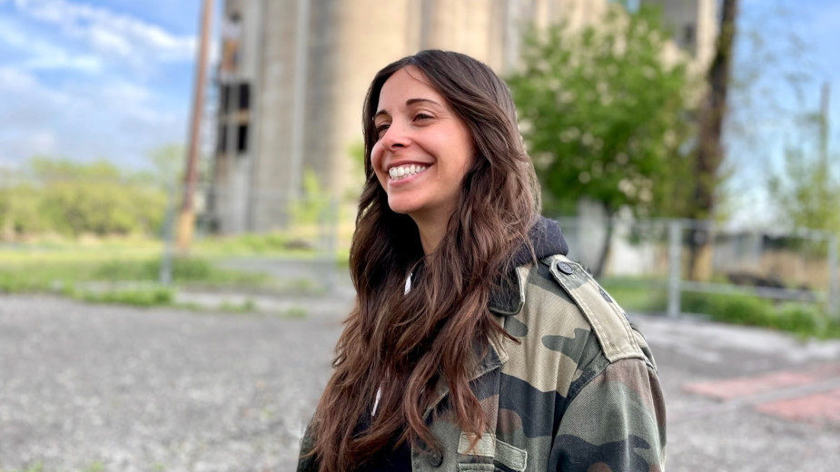A young woman with long brown hair wearing a camouflage jacket smiling outdoors with trees and a building in the background.