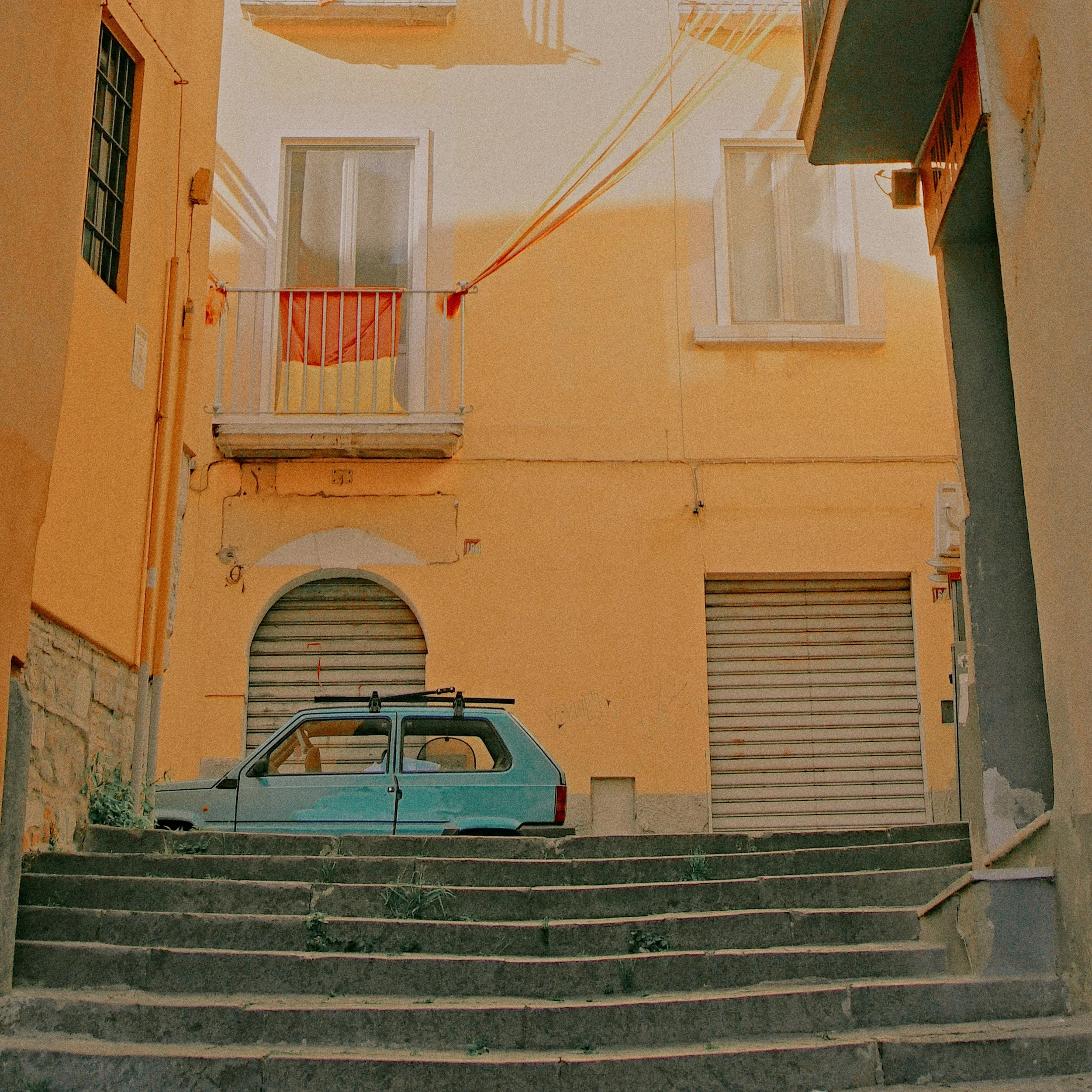 A small, vintage blue car parked on a set of stone stairs in front of a yellow building with two closed metal shutters and a balcony with a red towel hanging over the railing.