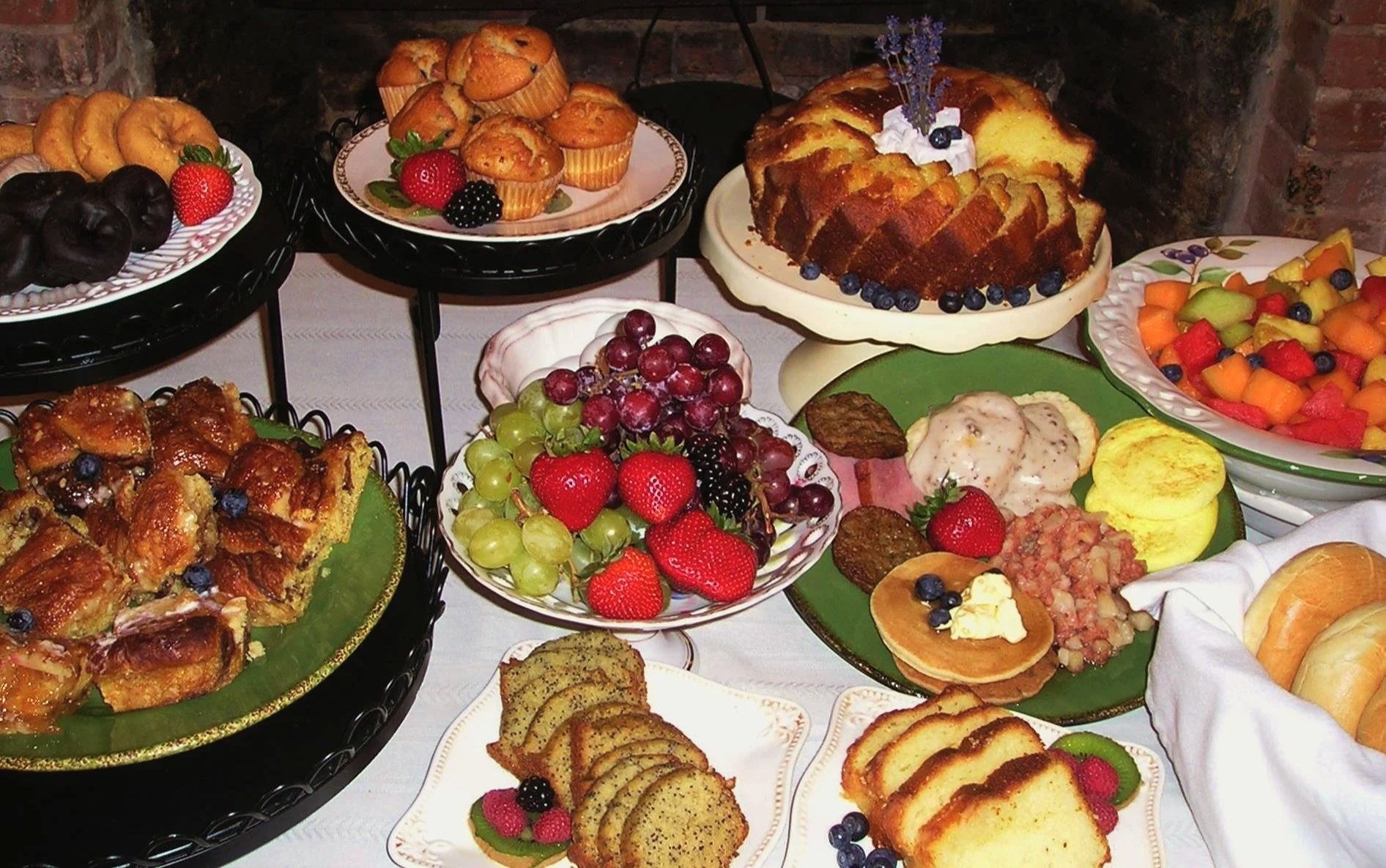 Assorted desserts and fresh fruits on a table, including cakes, muffins, fruit salad, grape bowls, and bread rolls.