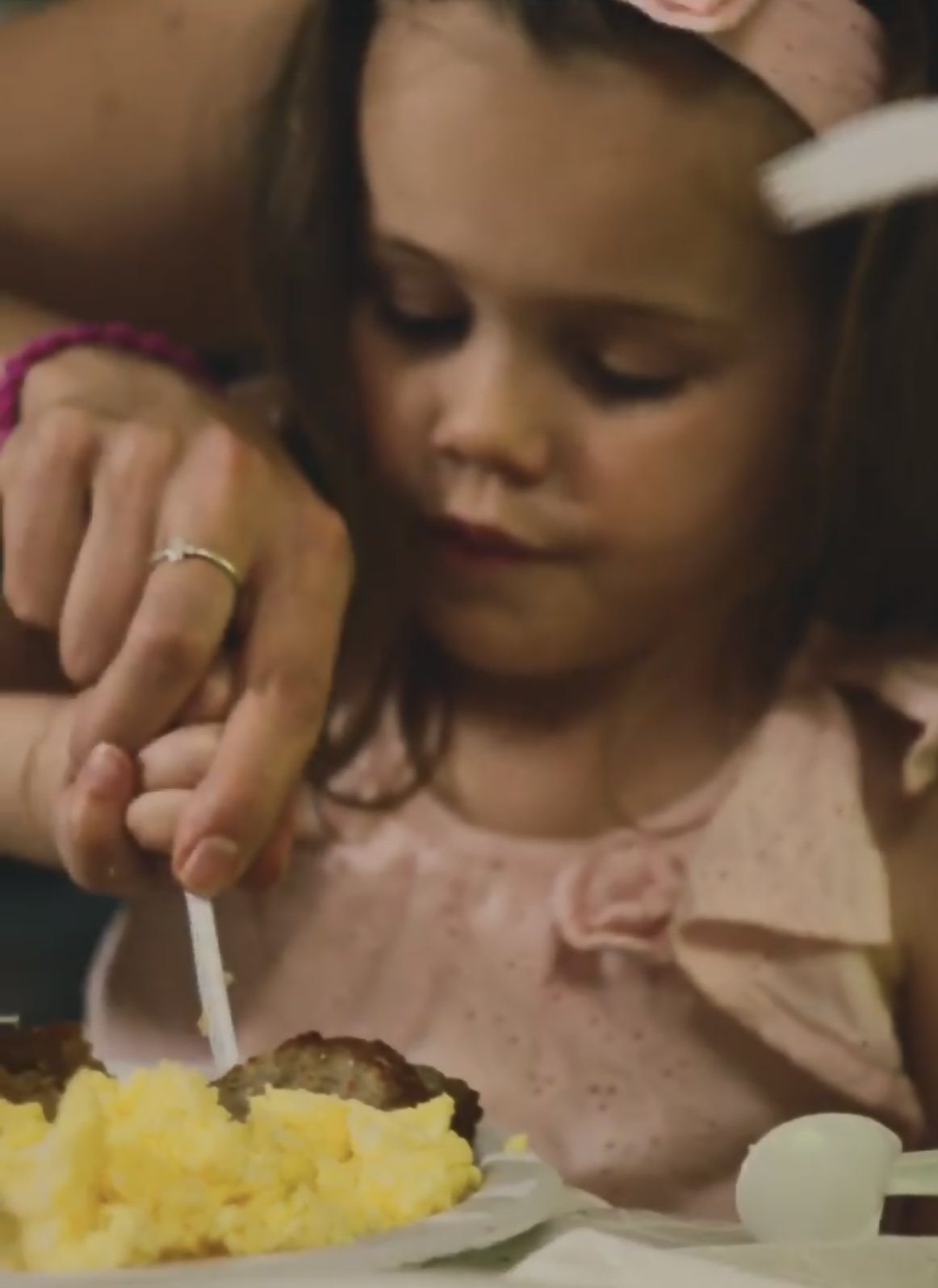 A young girl with dark hair and closed eyes being fed eggs and sausage by an adult woman, holding a fork, in a close-up view at a meal.