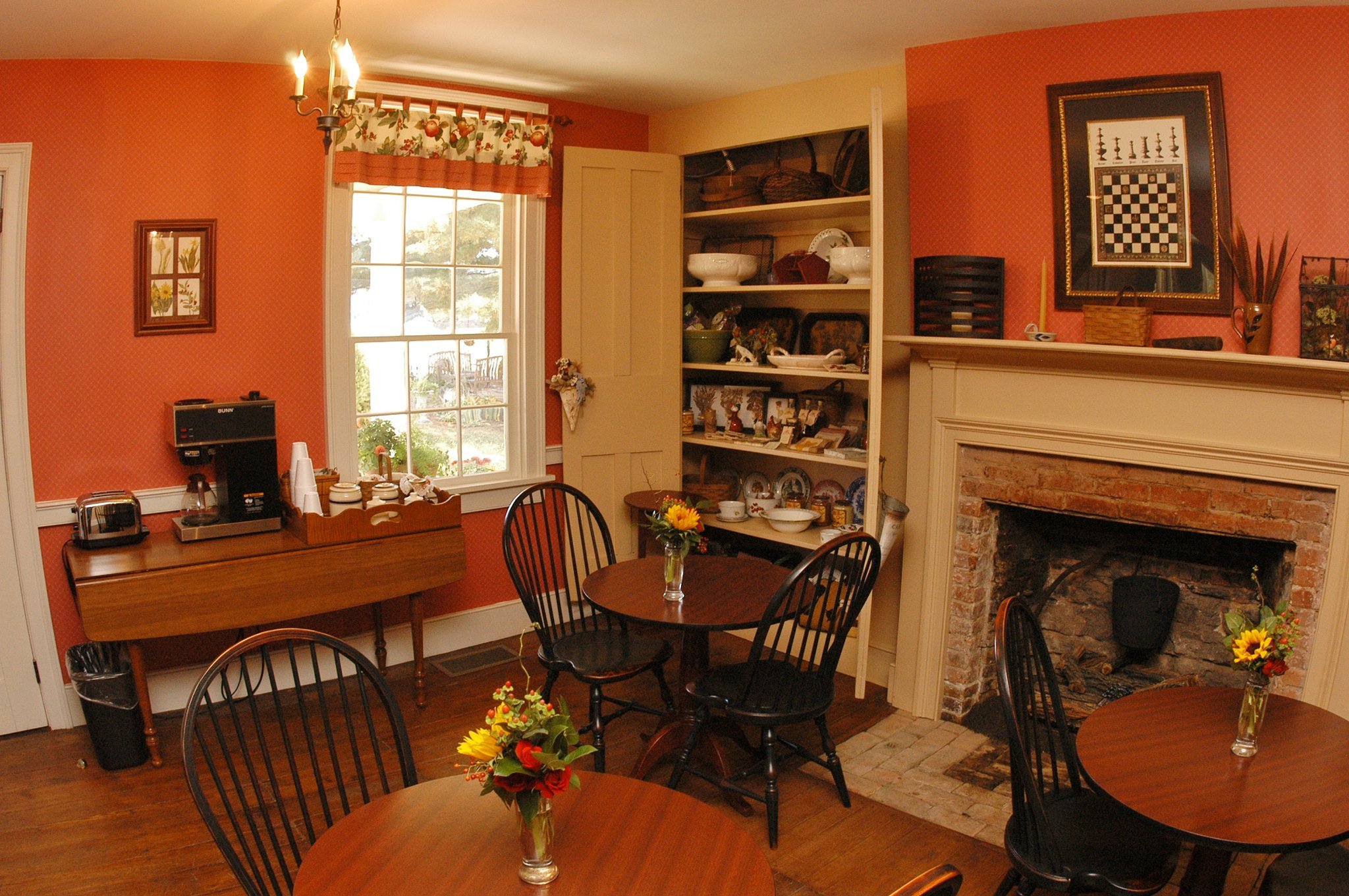 Cozy dining room with orange wallpaper, a fireplace, and wooden furniture. There are three round tables with flower vases, a china cabinet, and a coffee station. A large window with floral curtains lets in natural light.