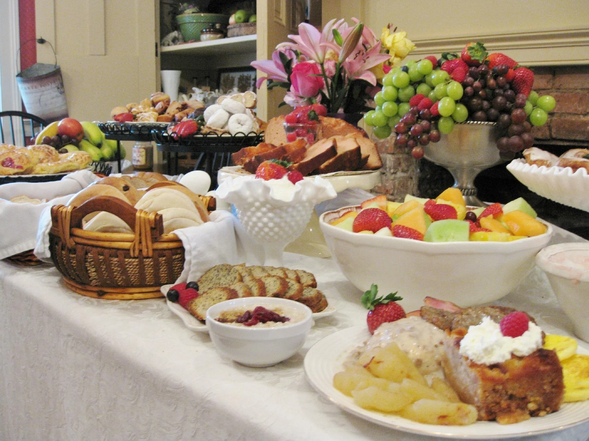 A table filled with assorted desserts, fruits, and baked goods set for a celebration, with a fruit centerpiece and flowers in the background.