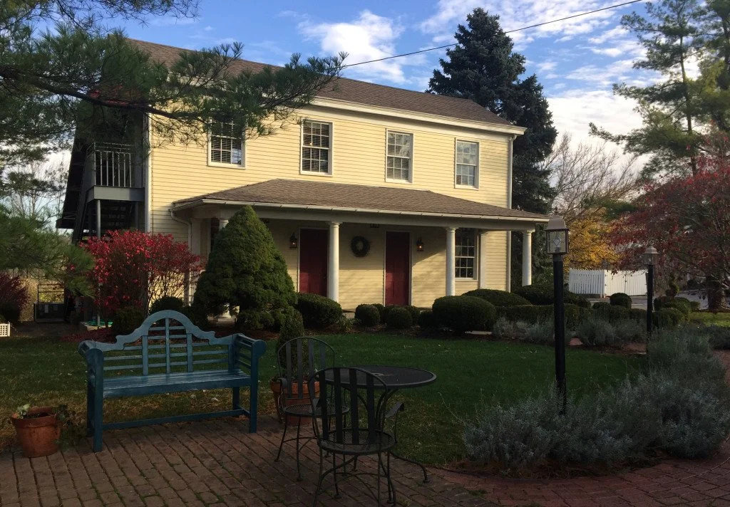 Yellow two-story house with white trim, a front porch, and red doors, surrounded by landscaped garden with bushes, trees, a blue bench, a small black table and chairs, a lamppost, and a brick pathway in a suburban neighborhood on a clear day.