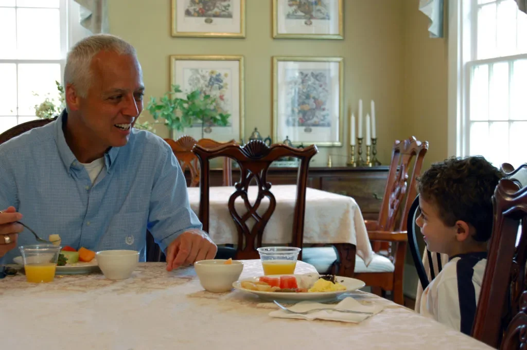 An older man and a young boy sitting at a dining table, smiling and having a conversation during breakfast, with plates of fruit, juice, and bowls on the table in a bright, decorated dining room.