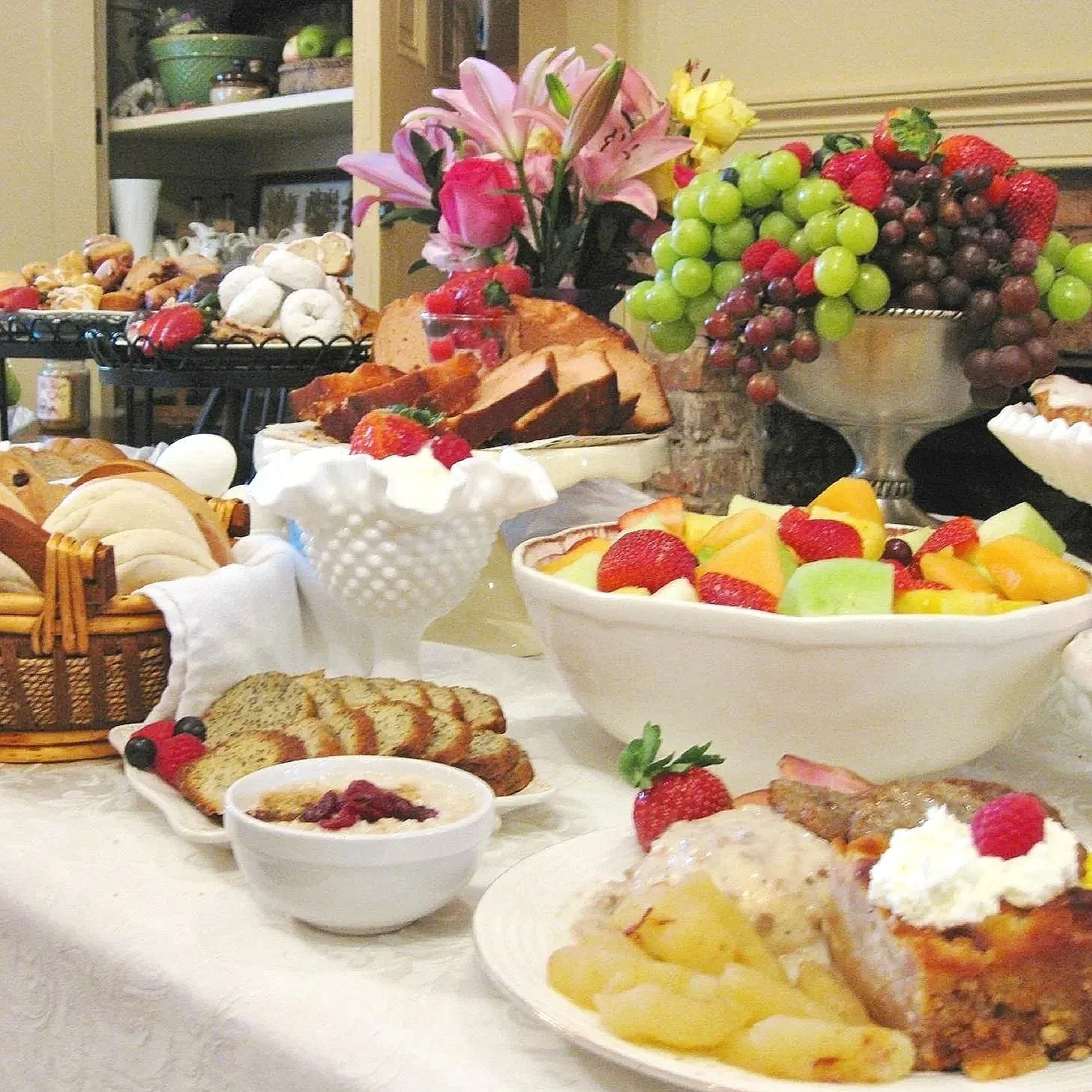 A table of various breakfast foods, including fresh fruit, sliced bread, pastries, yogurt, and a bowl of fruit salad, with flowers in the background.