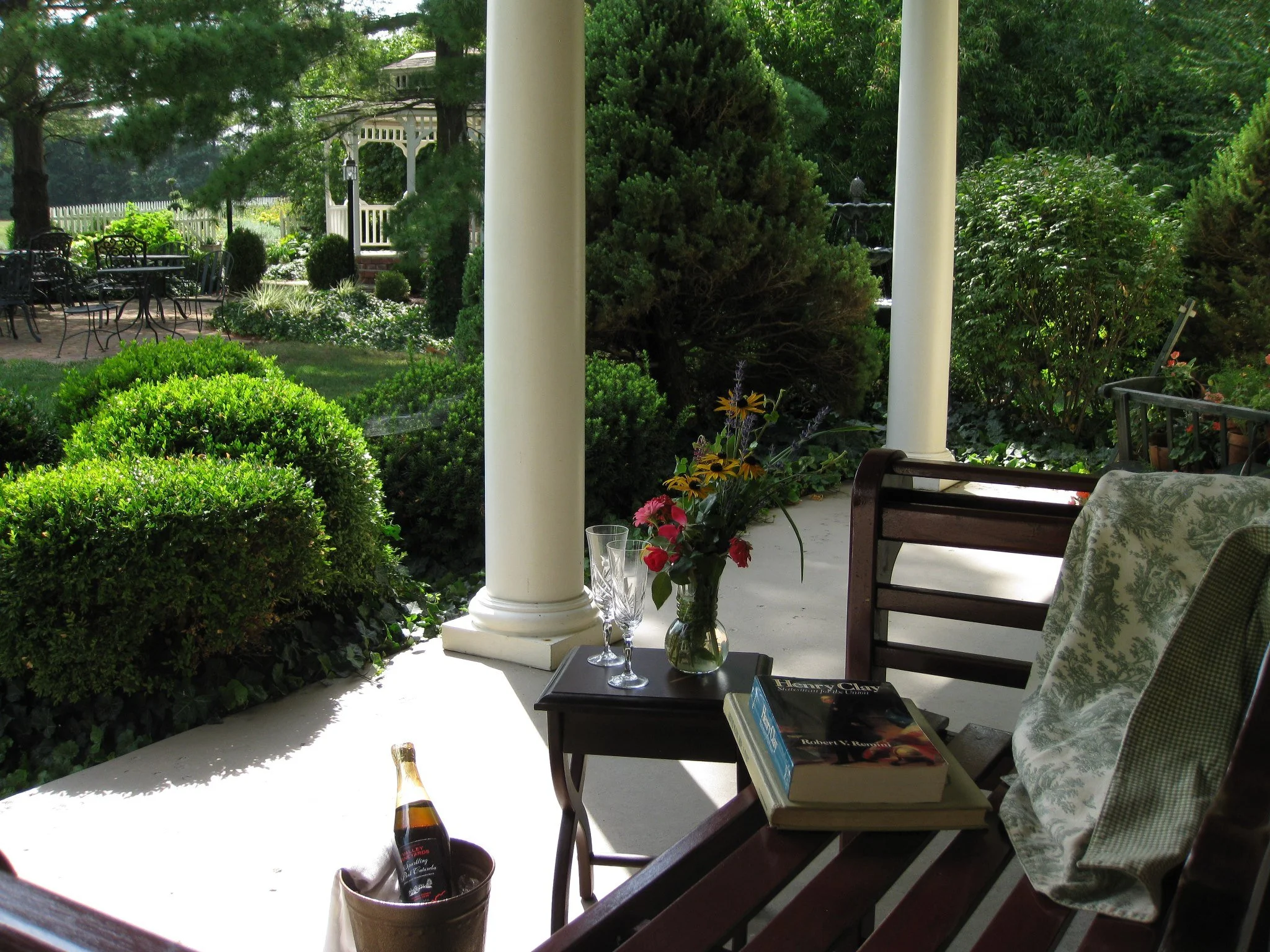 Porch with a wooden bench, a small table with a flower vase, books, and glasses, overlooking a lush garden with green bushes and trees.