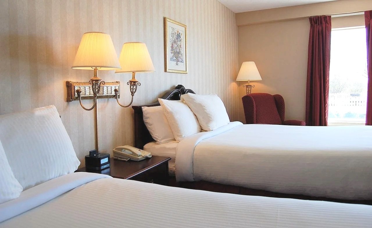 Two neatly made beds with white bedding in a hotel room, with a wooden nightstand between them holding a telephone and alarm clock, and two lamps mounted on the wall above. An armchair and a window with red curtains are visible in the background.