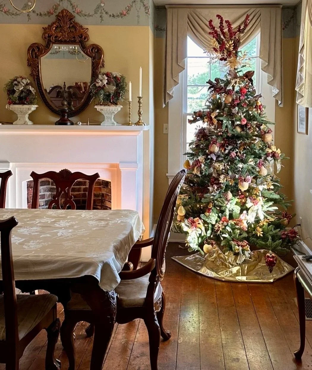 Decorated Christmas tree with ornaments near a window in a dining room with a wooden table and carved chairs, decorated festive garlands and candles on a mantelpiece.