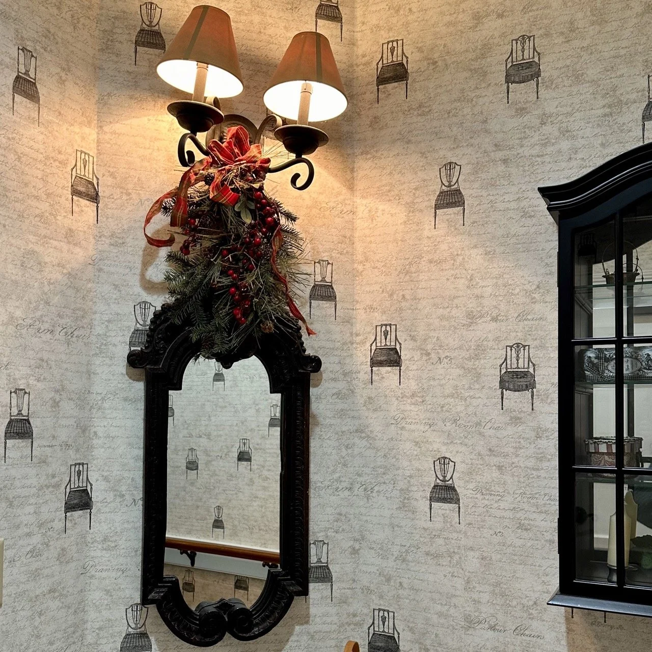 Decorative wall scene with a black carved mirror, wall-mounted light fixture with two lampshades, and Christmas greenery with red berries, ribbons, and bows. A black display cabinet is partially visible on the right.