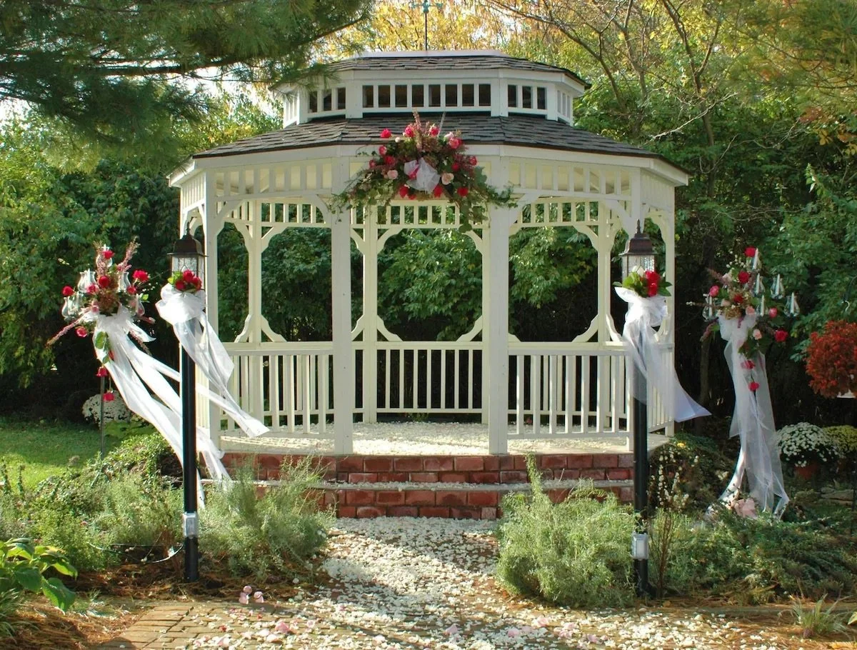 A white gazebo decorated with flowers and ribbons, set up for a wedding or special event in a garden with trees and plants.