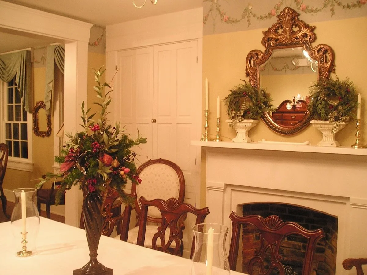 Elegant dining room with a floral arrangement on the table, a fireplace with a mirror above it decorated with greenery, and candles on the mantel