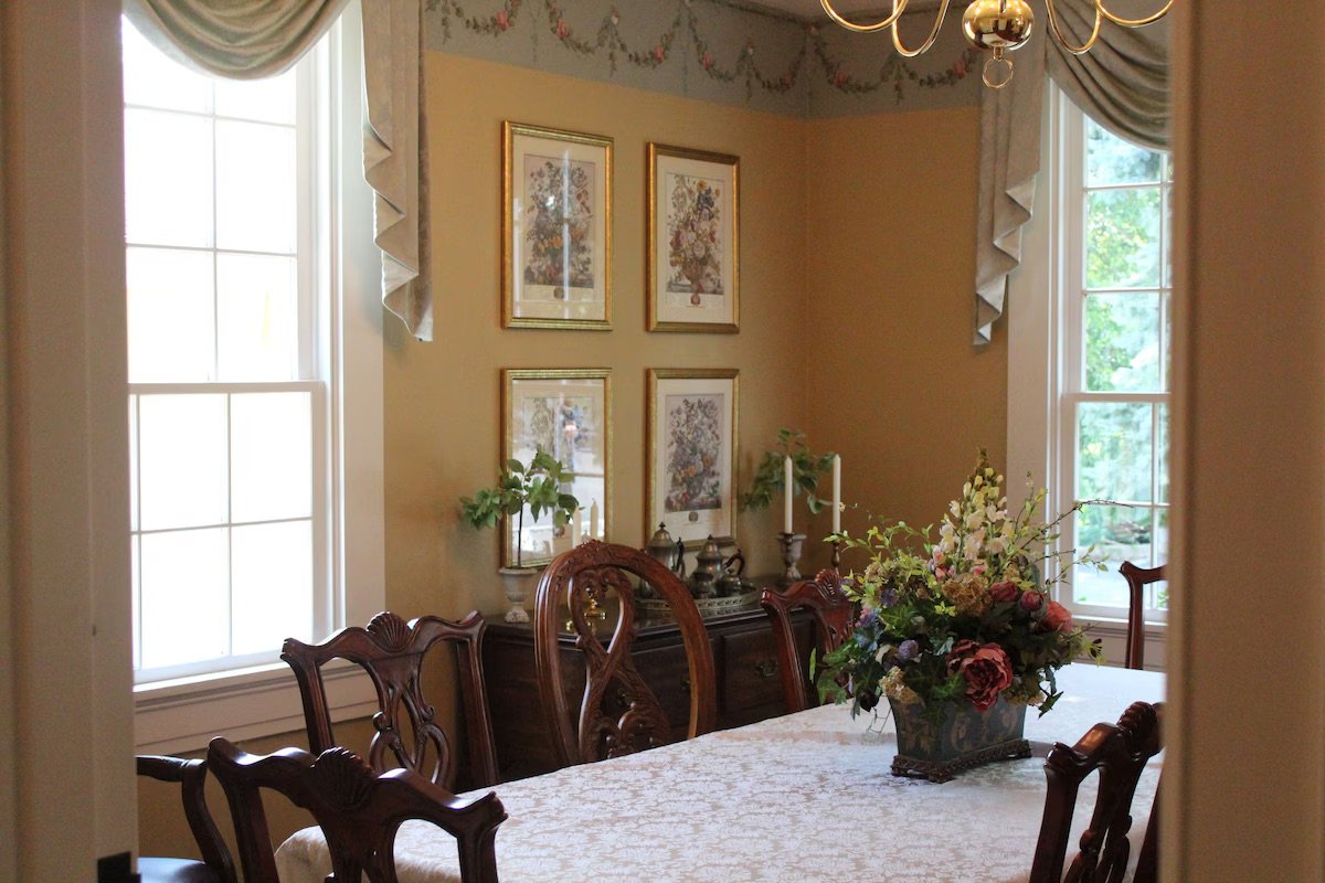 A dining room with a table covered with a white patterned tablecloth, a flower arrangement centerpiece, a china cabinet with decorative items, framed artwork on the wall, and two large windows with curtains letting in natural light.