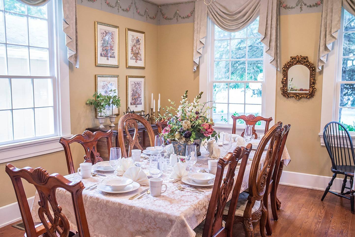 A dining room with a long table set for a formal meal, decorated with a floral centerpiece, candles, and fine china. The room has large windows with valances, framed artwork, and a gold-framed mirror, with hardwood flooring.