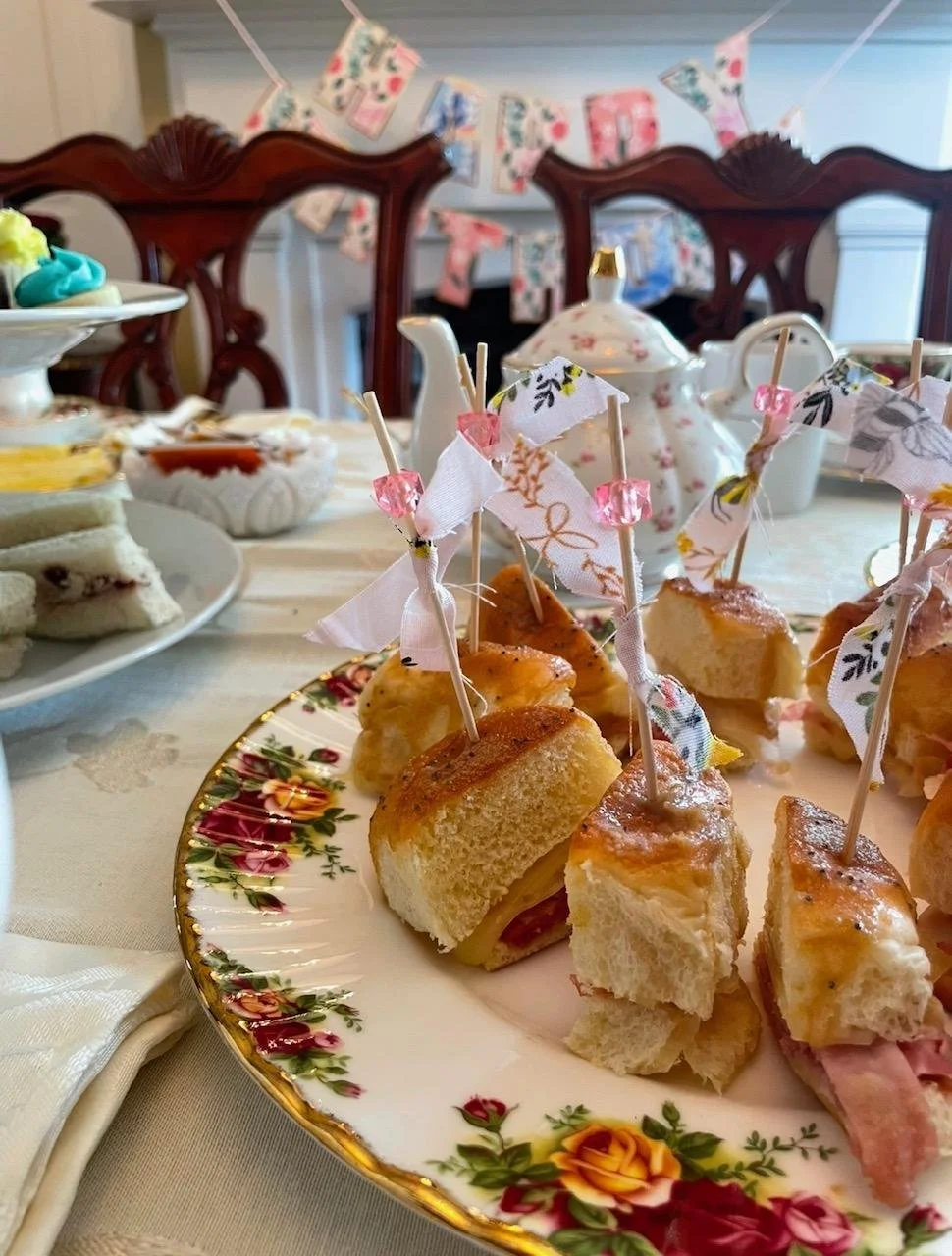 A fancy floral cake plate with small mini sandwiches and tea cakes, decorated with small fabric flags and pink beads. In the background, a teapot with floral pattern, a tiered tray with cupcakes, and a table with a tea party setup; a 'Happy Birthday'