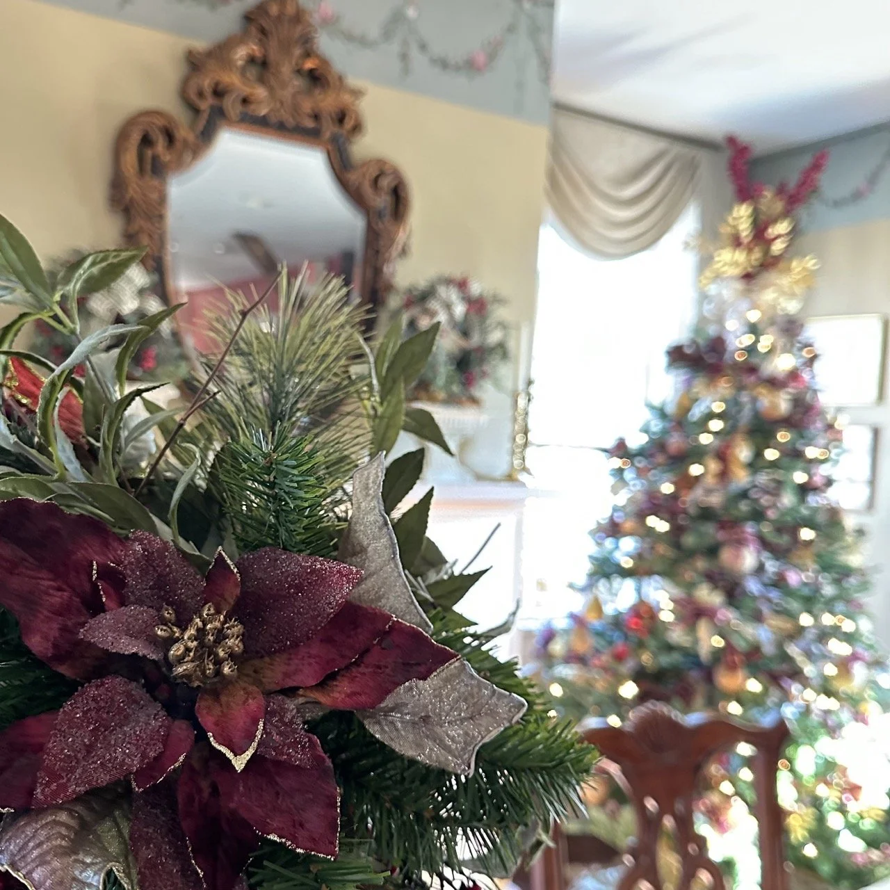 Close-up of a Christmas floral arrangement with poinsettias and greenery, with a decorated Christmas tree and festive decorations in the blurred background.