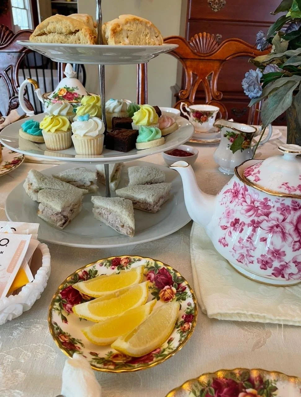 Tea set on a table with a three-tiered tray of assorted desserts, including finger sandwiches, cupcakes, brownies, and lemon wedges, with floral china teapot and cups, and decorative china on a lace tablecloth.