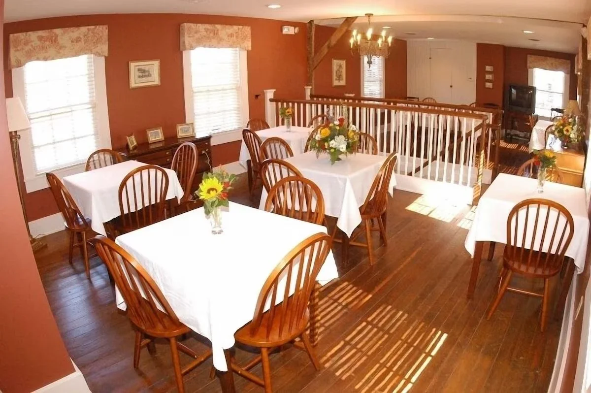 A dining area with four tables covered in white tablecloths, each decorated with a flower arrangement. The room has wooden floors, brown walls, and is lit by natural light from windows and a chandelier. There is a small railing overlooking a lower le
