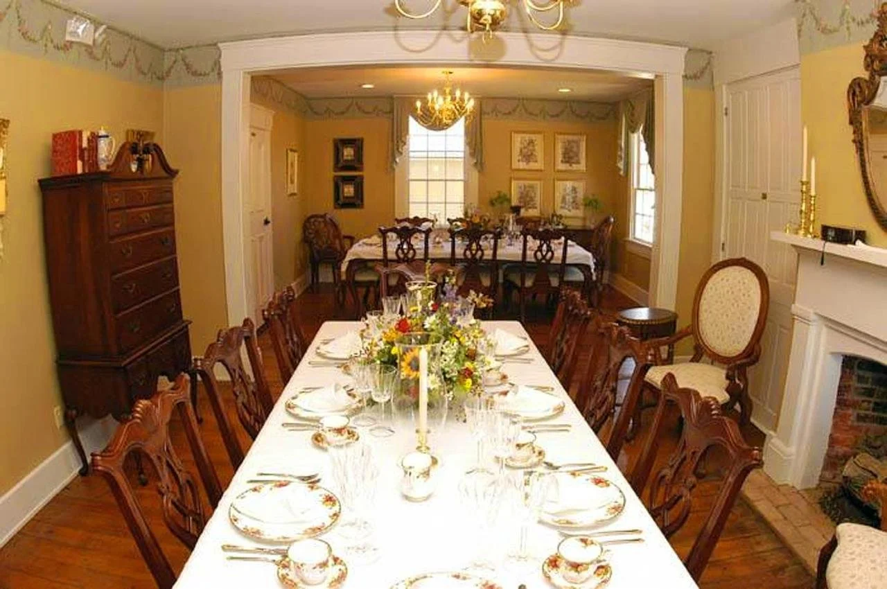 A dining room with a long table set for a formal meal, featuring floral centerpiece, crystal glasses, and plated dinnerware. The room has yellow walls, hardwood flooring, and decorative window treatments. A chandelier hangs above the table, and there