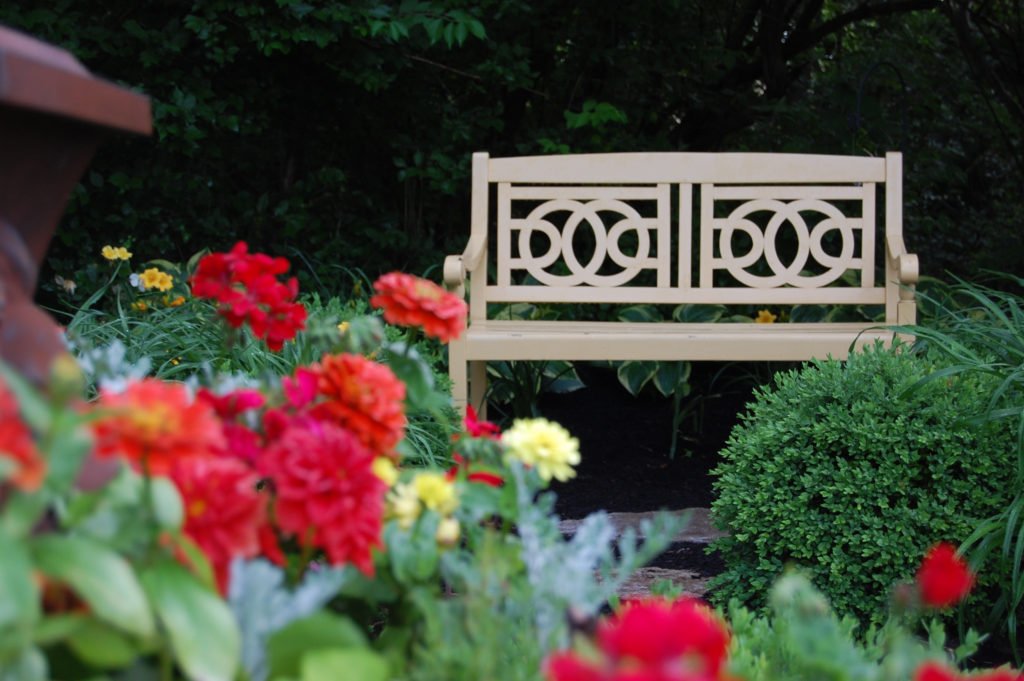 A cream-colored decorative wooden bench in a garden surrounded by colorful flowers and green bushes.