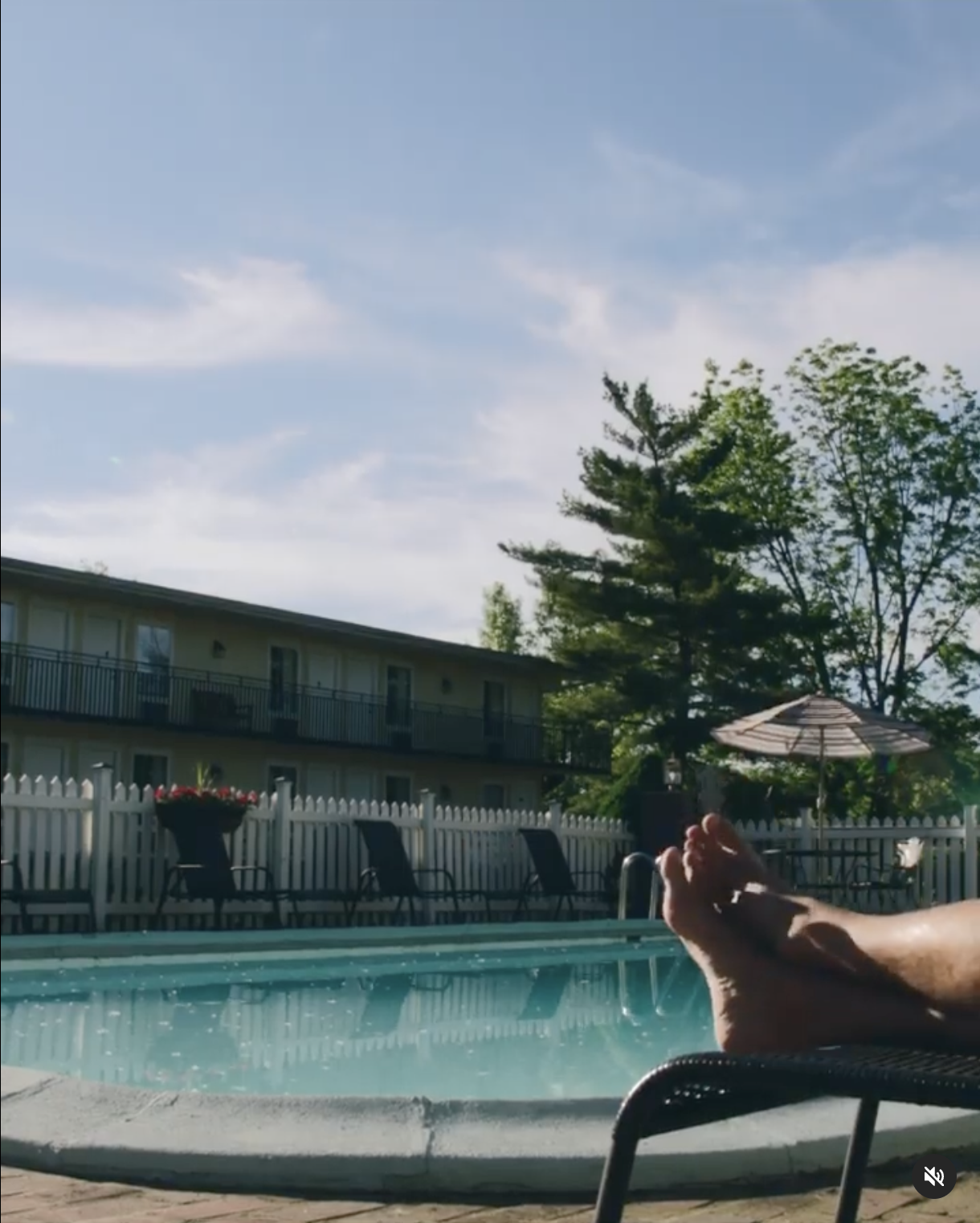 Person relaxing on a lounge chair by a swimming pool, with buildings, trees, and blue sky in the background.