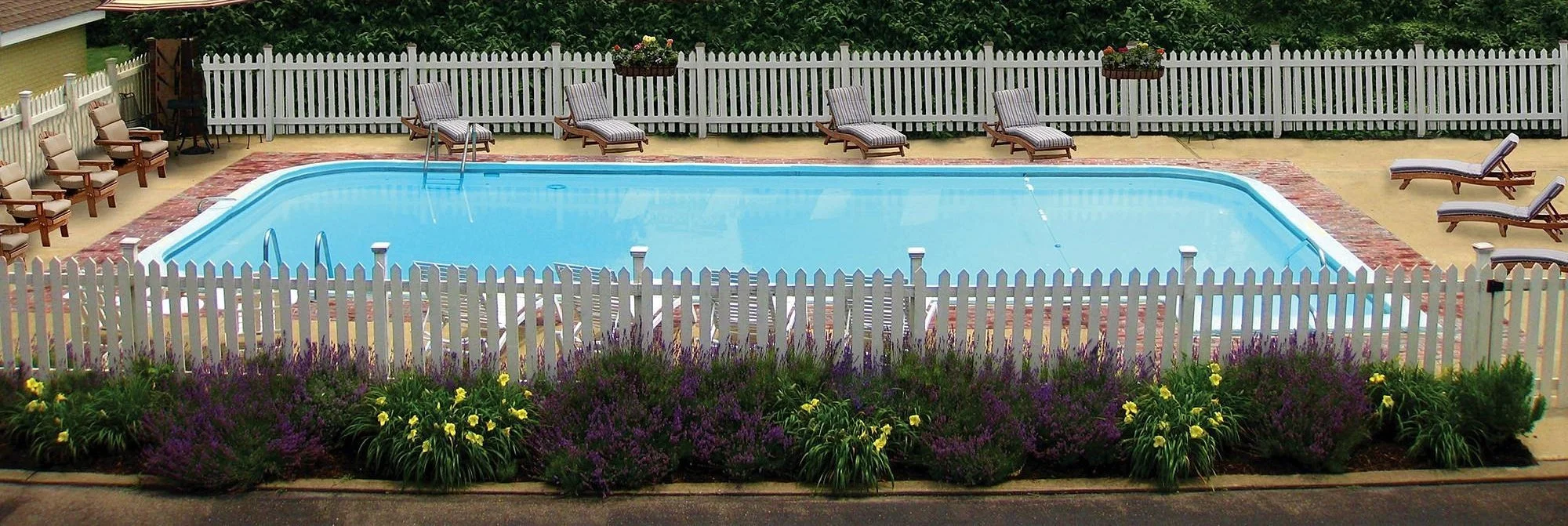 A backyard swimming pool with lounge chairs, surrounded by a white picket fence and flower beds.