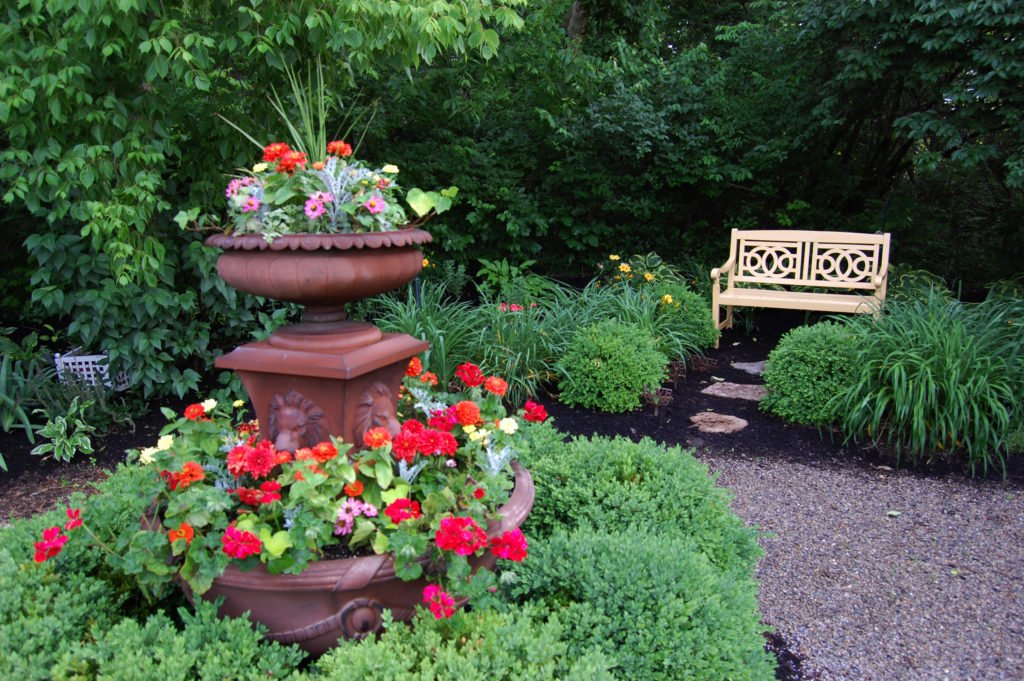 A garden scene featuring a two-tiered terra cotta fountain with colorful flowers, surrounded by green bushes and plants. In the background, there is a wooden bench among lush foliage.