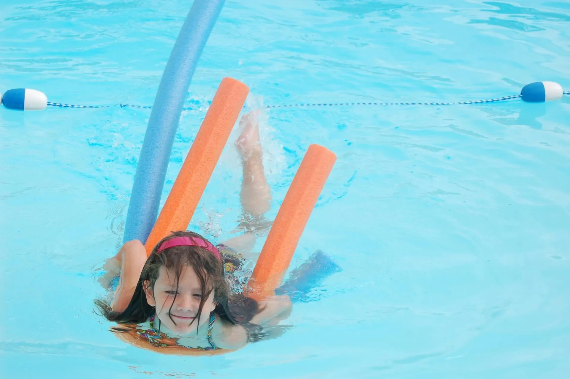 Young girl with wet hair swimming in a pool, wearing a pink swimsuit and holding onto pool noodles with blue and orange colors.