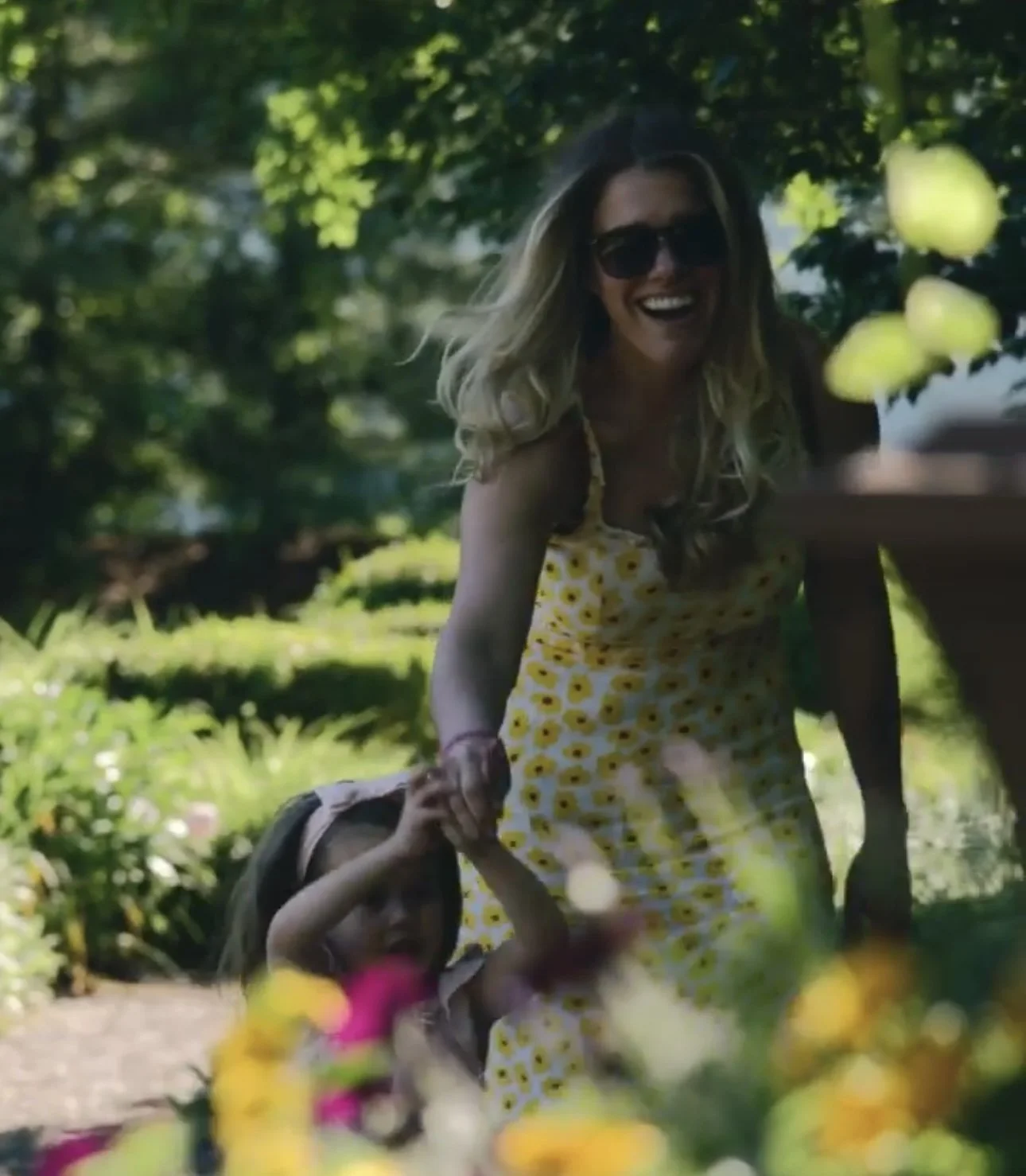 A woman and a young girl walking outdoors in a sunny garden or park, smiling and holding hands. The woman is wearing sunglasses and a yellow dress with a floral pattern. The girl has long hair, and the background is lush with green trees and colorful