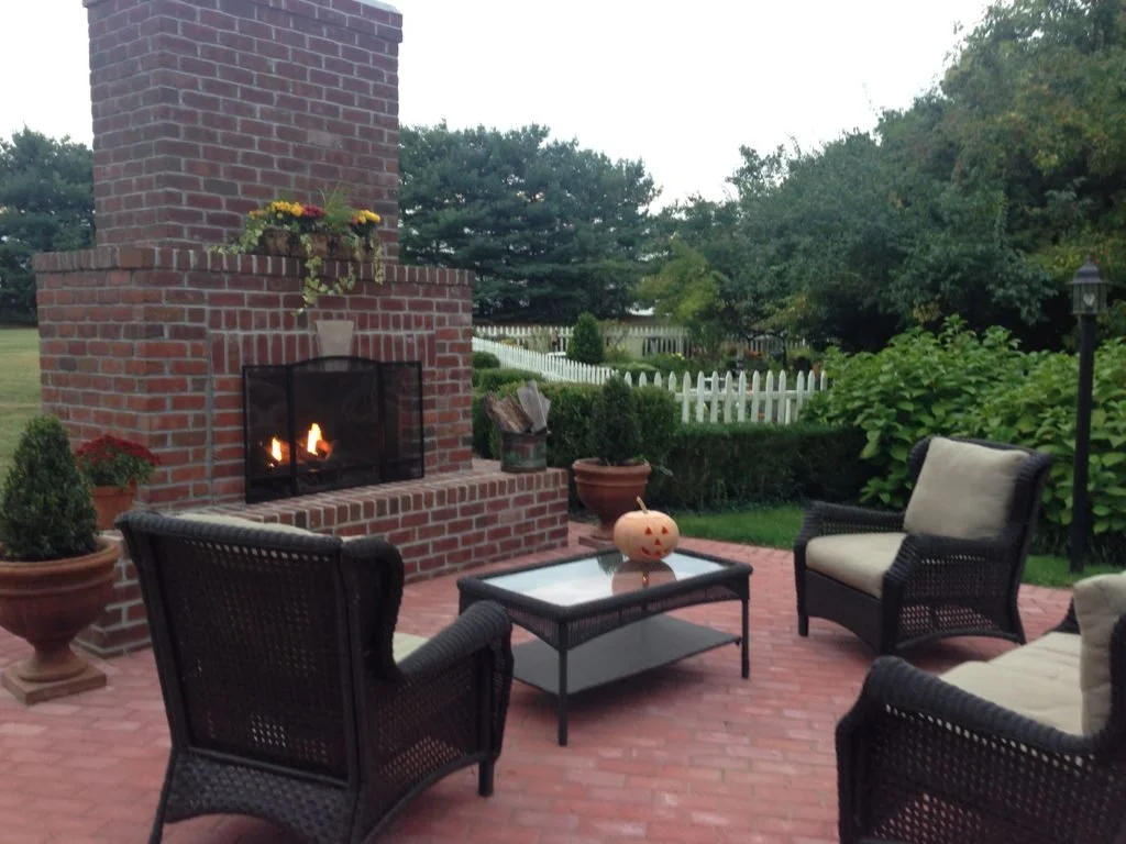 Outdoor patio with a brick fireplace, black wicker chairs with beige cushions, a glass-topped table with a carved pumpkin, potted plants, and a white picket fence in the background.
