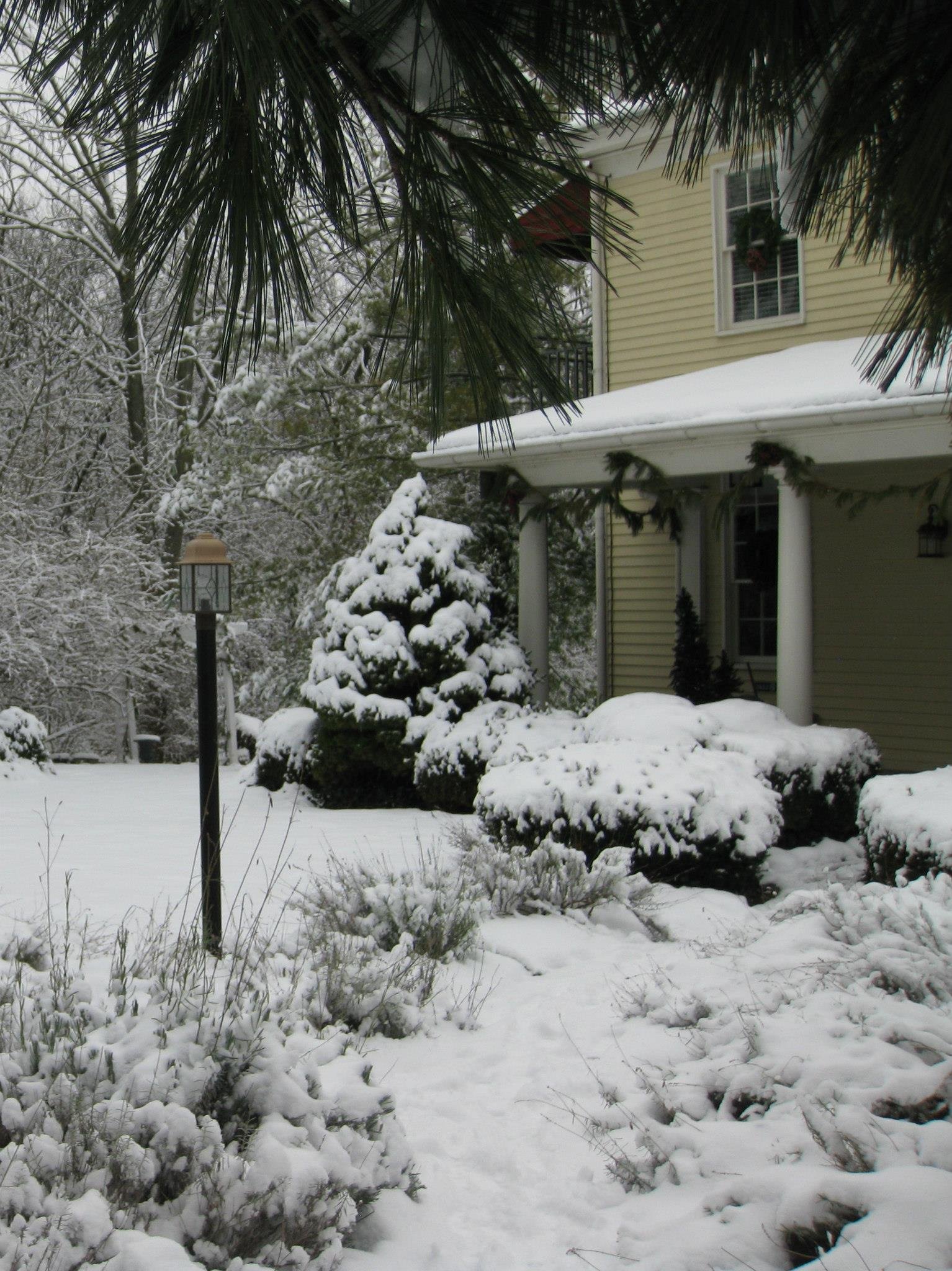 Snow-covered garden and house with white siding, porch columns, and decorative garlands, framed by tree branches and a lamp post.
