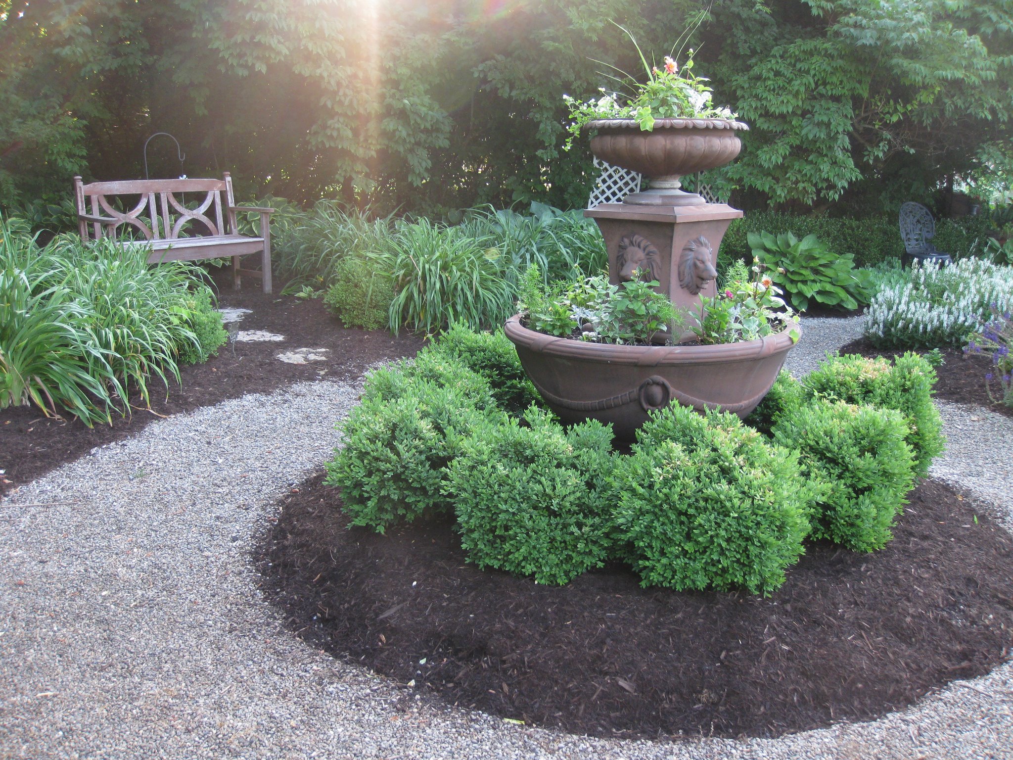 A garden scene with a central decorative fountain surrounded by green bushes and flowering plants, with a wooden bench on the left and metal chairs on the right, under trees with sunlight filtering through.
