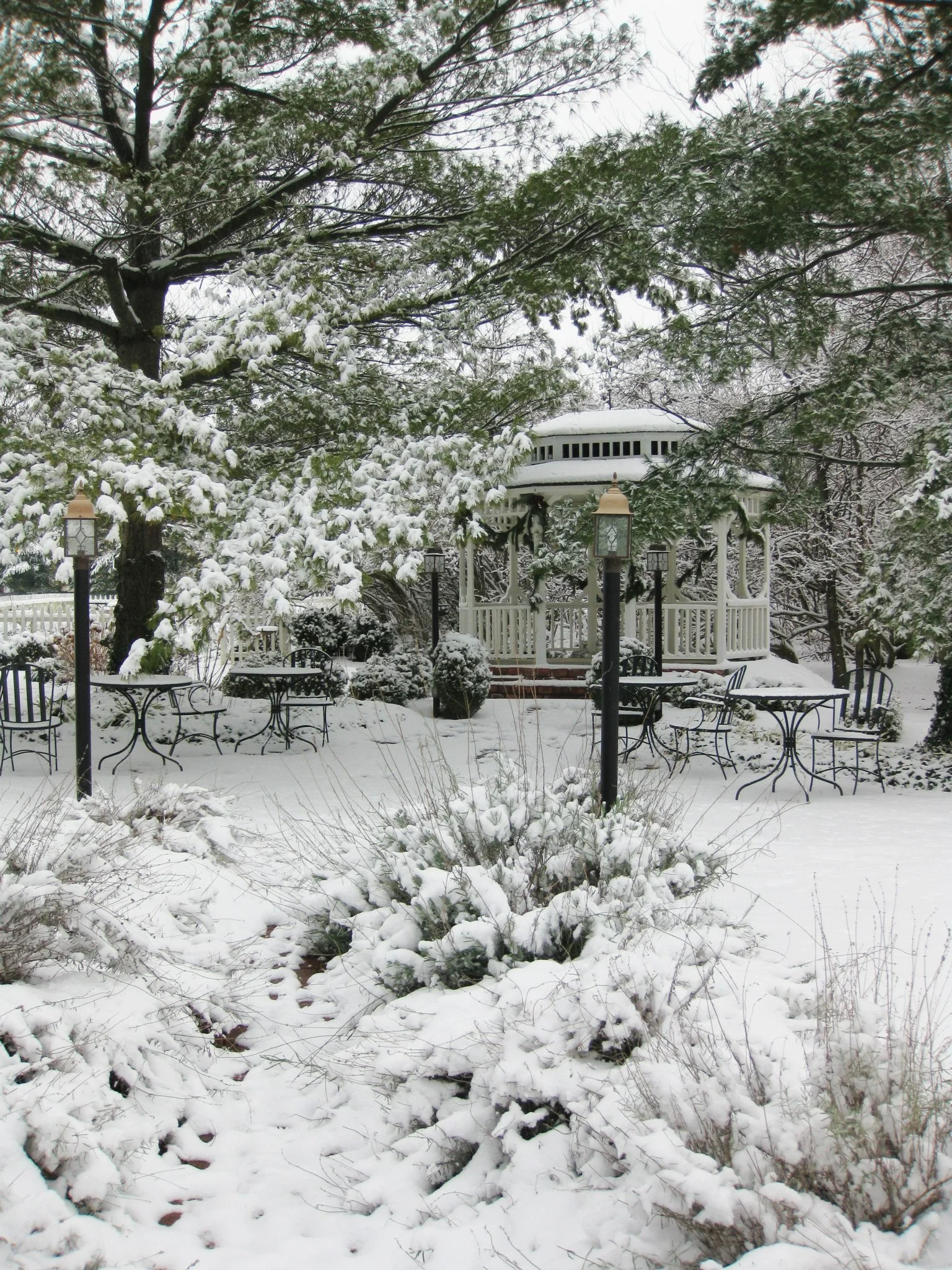 Snow-covered garden with trees, a white gazebo, black metal chairs, and street lamps.