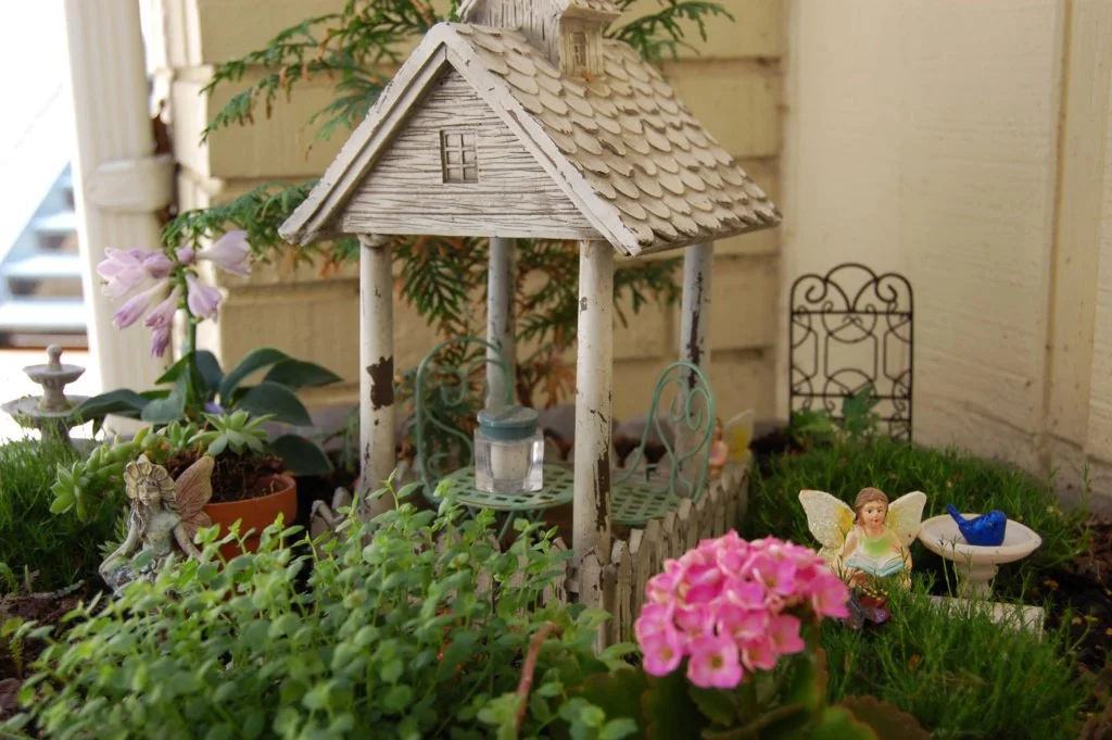 A small, decorative garden scene featuring a miniature white wooden gazebo with a shingled roof, surrounded by potted plants, pink flowers, a fairy garden figurine, an angel figurine, and a tiny birdbath with a blue bird ornament.