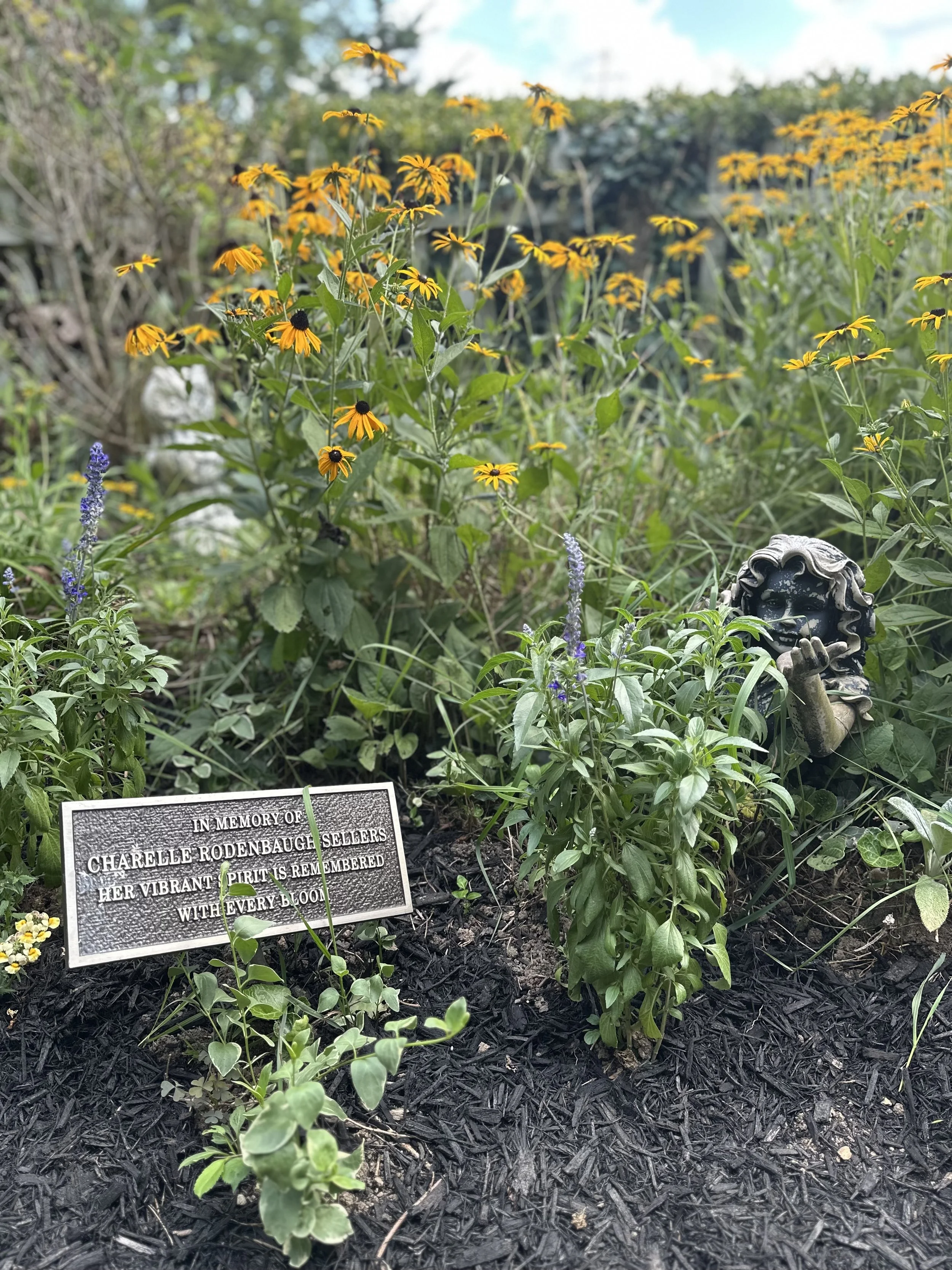 A garden bed with yellow and purple flowers, a memorial plaque in memory of Charelle Rodenbaugh, and a decorative garden statue resembling a smiling face with curly hair.