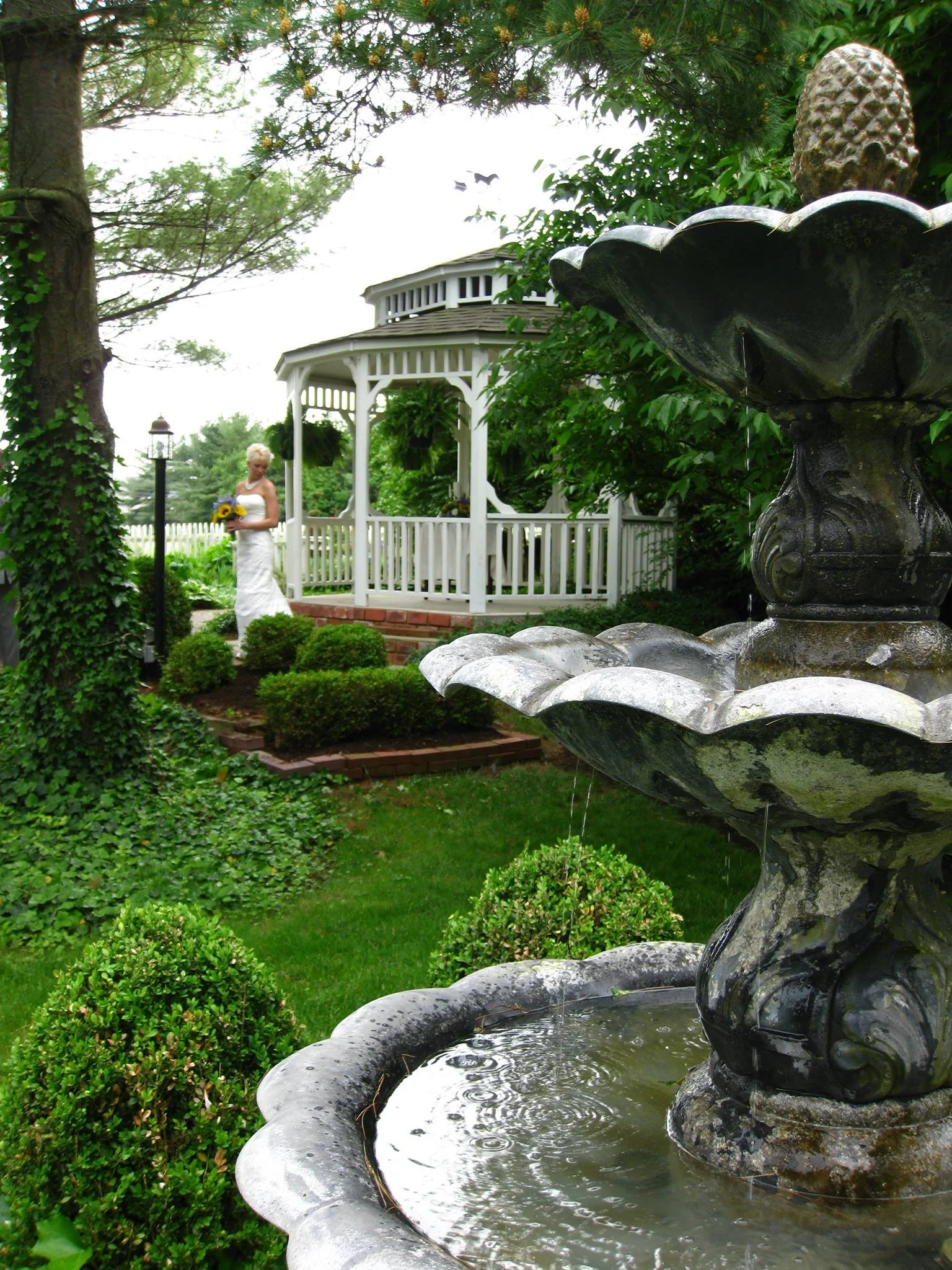 A bride in a white dress holding a bouquet of sunflowers standing near a white gazebo in a lush garden, with a stone fountain in the foreground.