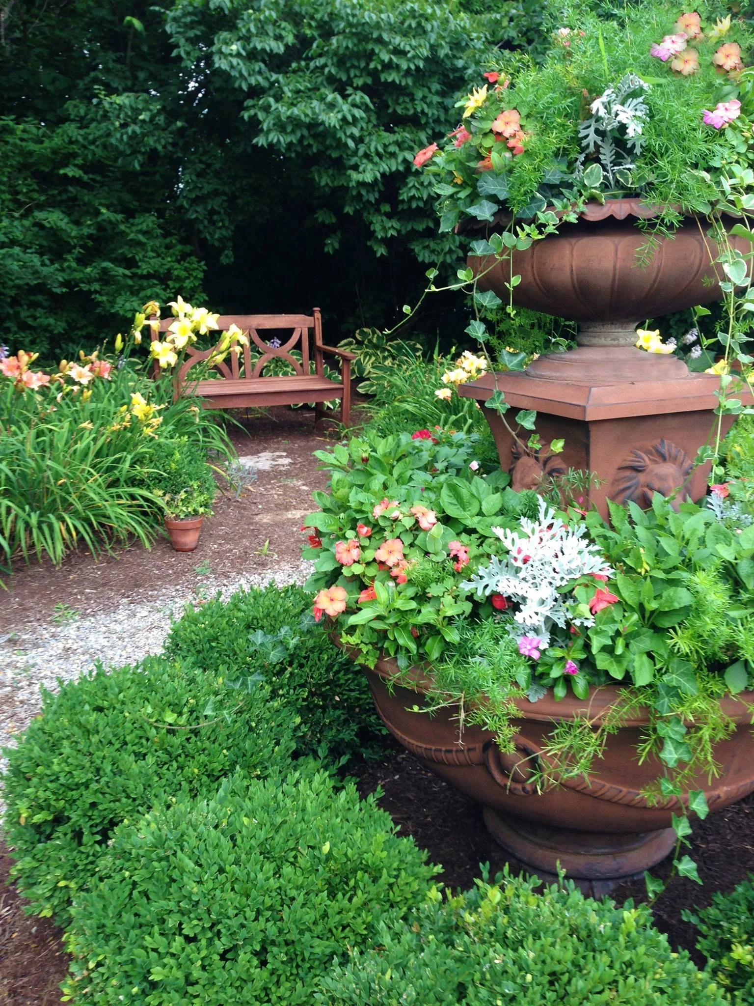 A garden scene with a large terracotta urn overflowing with pink, yellow, and white flowers. A wooden bench is in the background, surrounded by lush greenery and various plants.