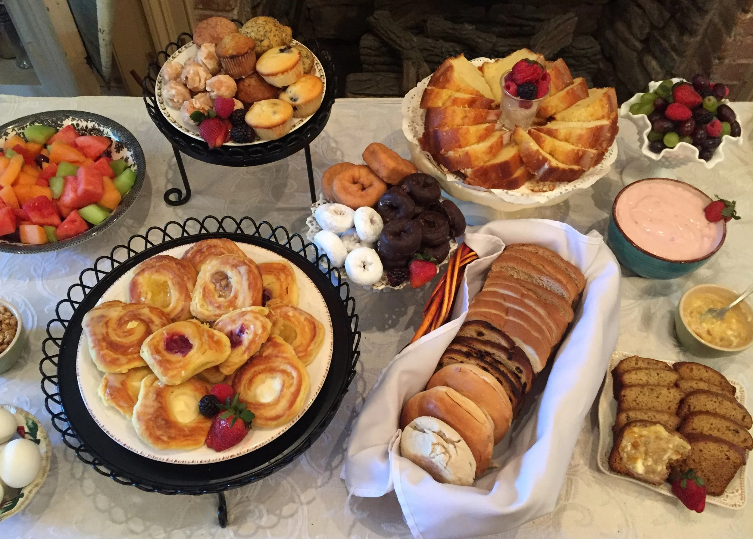A table with an assortment of desserts and breakfast items, including fruit salad, muffins, donuts, a bundt cake with berries, assorted cookies, sliced bread, and bowls of yogurt and fruit.