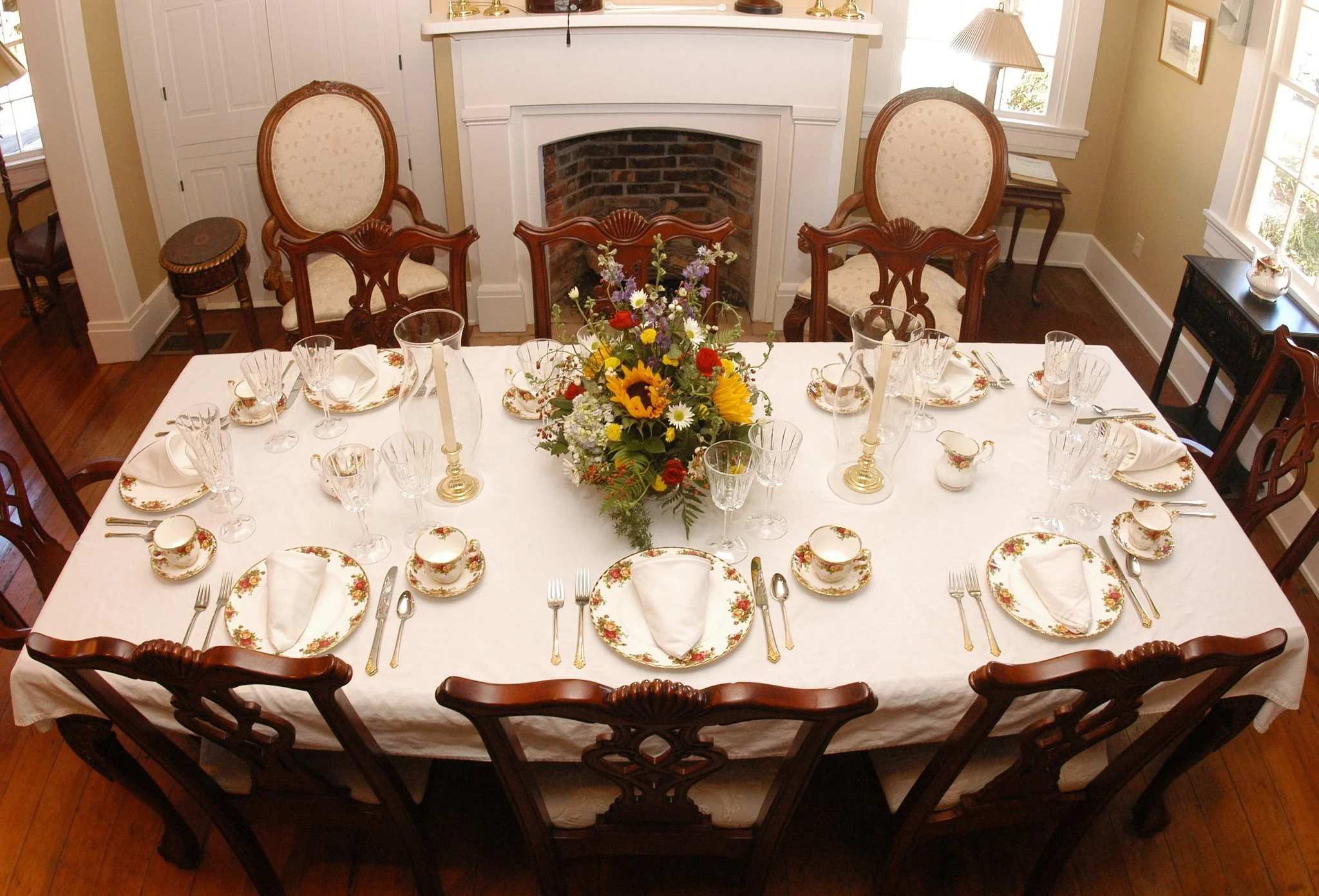 A dining table set for a meal in a room with hardwood floors, a brick fireplace, and windows. The table has a white tablecloth, floral china, white napkins, and glassware. A large floral centerpiece with sunflowers, daisies, and other flowers sits in the middle. There are four candles in brass holders on the table.