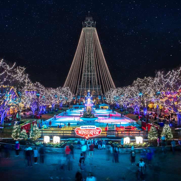 A festive ice skating rink decorated with Christmas lights at night, with a large illuminated Christmas tree at center, surrounded by trees with colorful lights, and a tall Christmas star at the top of a structure resembling a Christmas tree. A sign reads "Kings Island Winterfest."
